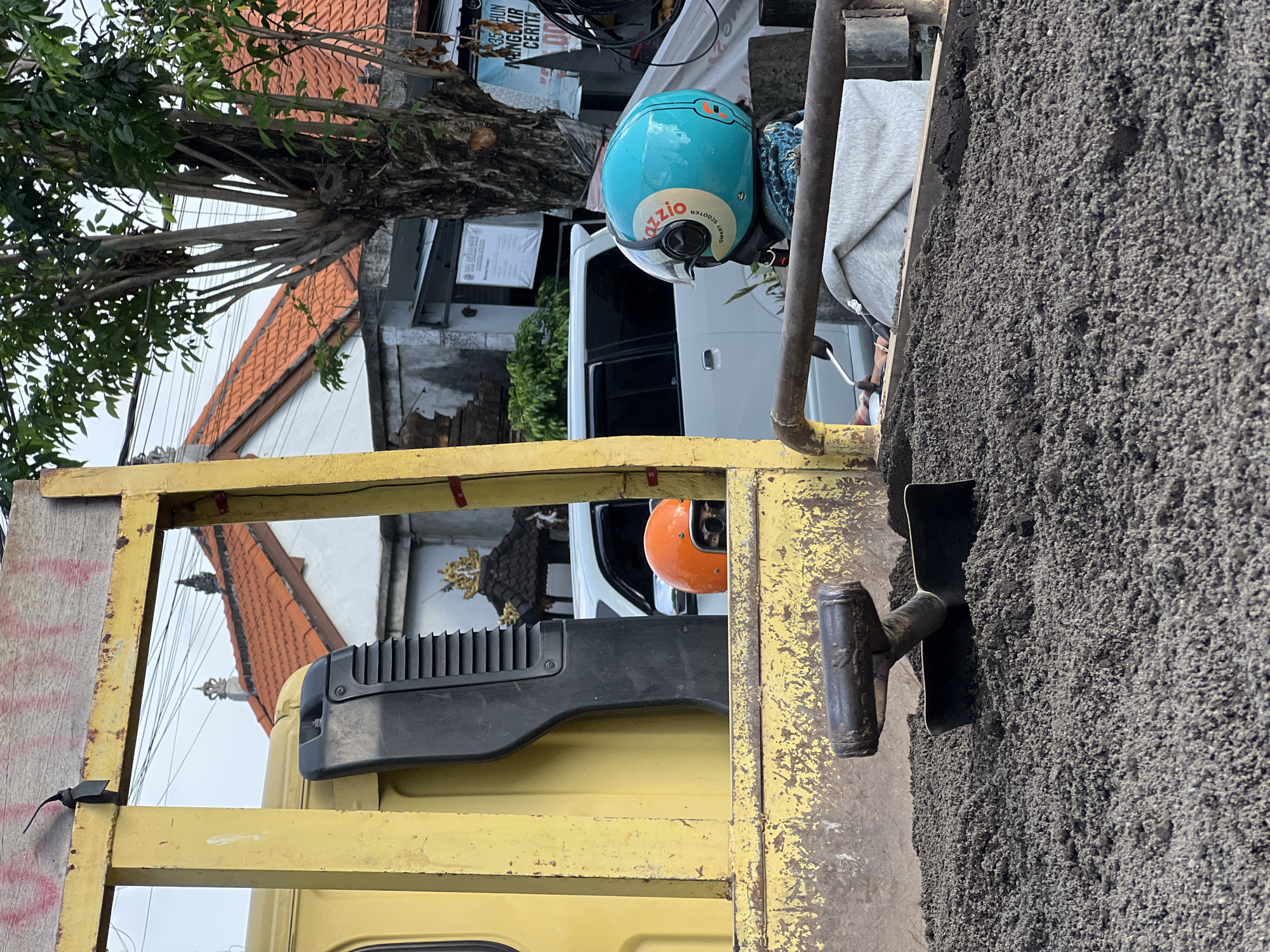 The image shows the contents of a truck bed, filled with a dark, granular material, likely gravel or sand, located in Denpasar Barat, Indonesia. A hand trowel with a dark handle is embedded in the material. A yellow metal framework of the truck bed is visible. In the background, partially obscured, are urban elements: a white van, a blue Azzio brand helmet resting on something, and the rooftops of buildings with orange tiles, characteristic of the region. A tree with dense foliage is also visible. The scene appears to be a delivery or construction site.