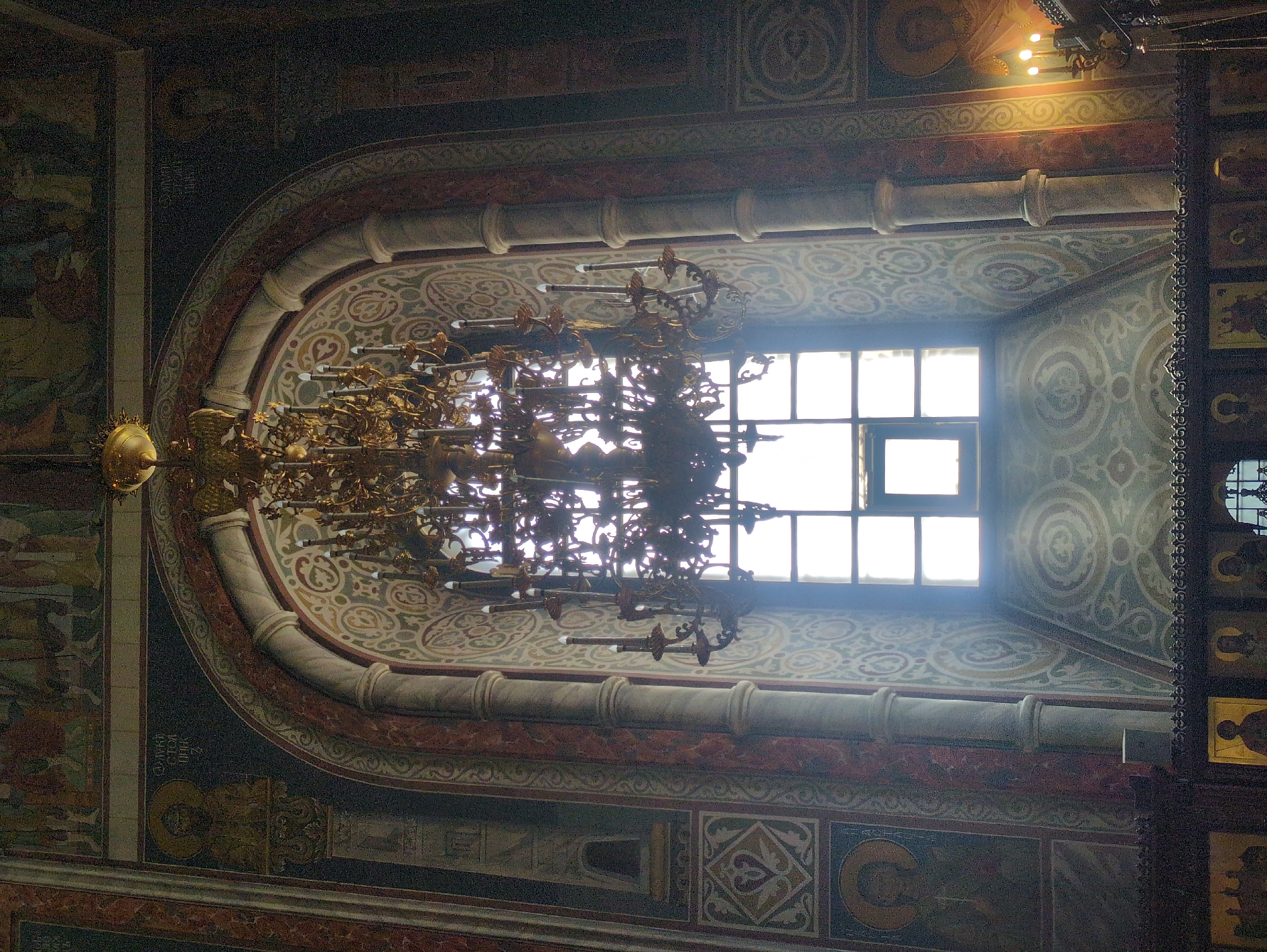 This is a photograph taken inside a church in Nizhny Novgorod, Russia. The focal point of the image is a large, ornate, golden chandelier with many candle-like lights, hanging in front of a tall, arched window. The window is divided into multiple panes and is brightly lit from behind, suggesting it is daytime. The walls of the church are richly decorated with frescoes and intricate patterns in muted colors. In the foreground, a dark wooden iconostasis with several icons is partially visible. The overall impression is one of historical and religious grandeur.