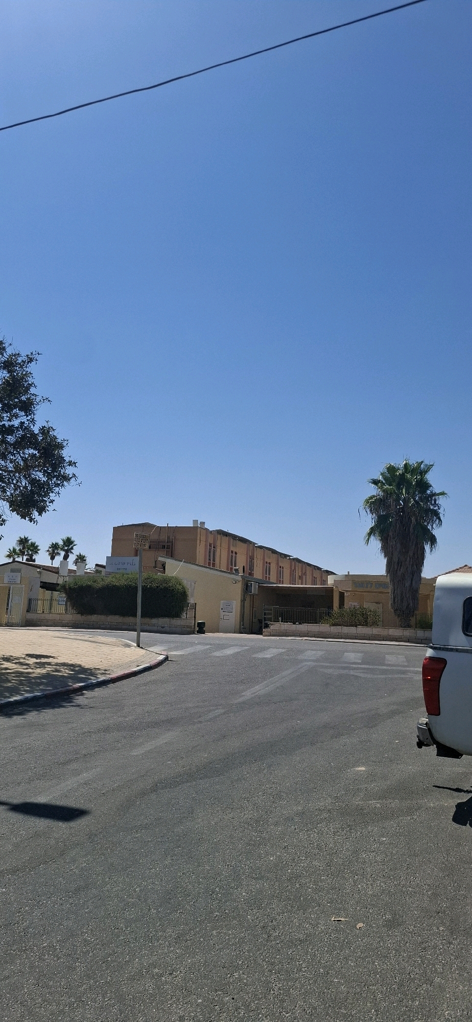 The image depicts a street scene in Yerocham, Israel, on a clear day. A dark power line is visible diagonally across the bright blue sky. In the middle ground, a long, tan-colored building with multiple stories and rows of windows stands prominently. In front of this building, there are several smaller, single-story structures. A tall, mature palm tree with drooping fronds is located to the right of the main building. To the left of the main building, a smaller tree with dark green foliage is visible. The foreground shows a paved road with white pedestrian crossing lines painted on it. A section of a white pickup truck is visible on the right side of the road, with its taillight and bumper in frame. A shadow, likely cast by a street light, stretches across the road from the left.