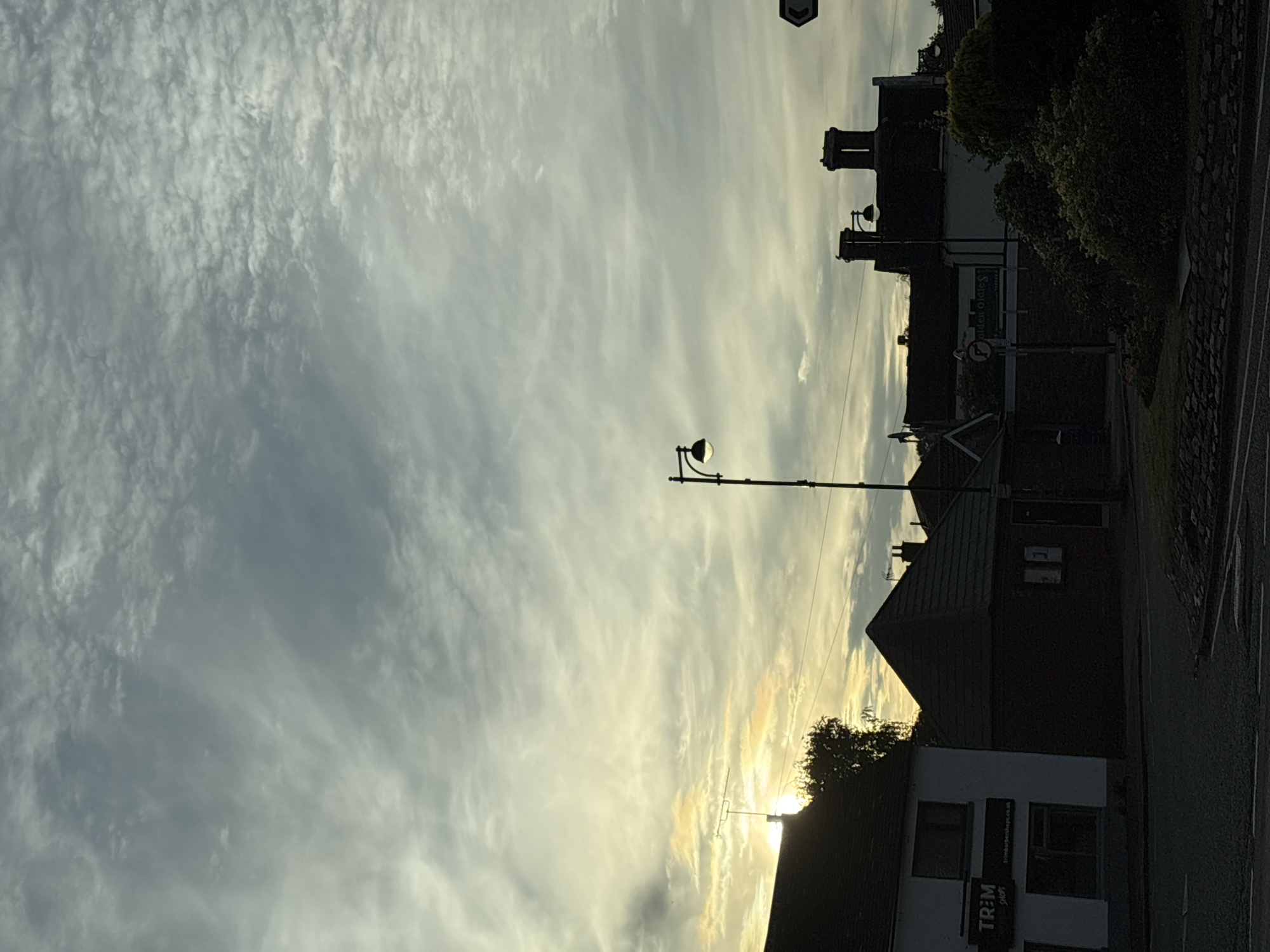 The image shows a street scene in South Staffordshire, United Kingdom, under a cloudy sky with the sun partially visible through the clouds, creating a soft, diffused light. On the left, a building with a dark roof and white facade displays a sign that reads "TRIM SHOPS" with "trimbarbershops.co.uk" below it. To the right of this building is a smaller structure with a dark, pyramid-shaped roof. Further in the background, several buildings with chimneys are visible, silhouetted against the sky. A street lamp with a globe is positioned between these buildings. In the foreground, the edge of a road with a stone curb is visible. The scene is dominated by the sky, which is filled with various cloud formations. There are no people visible in the image.