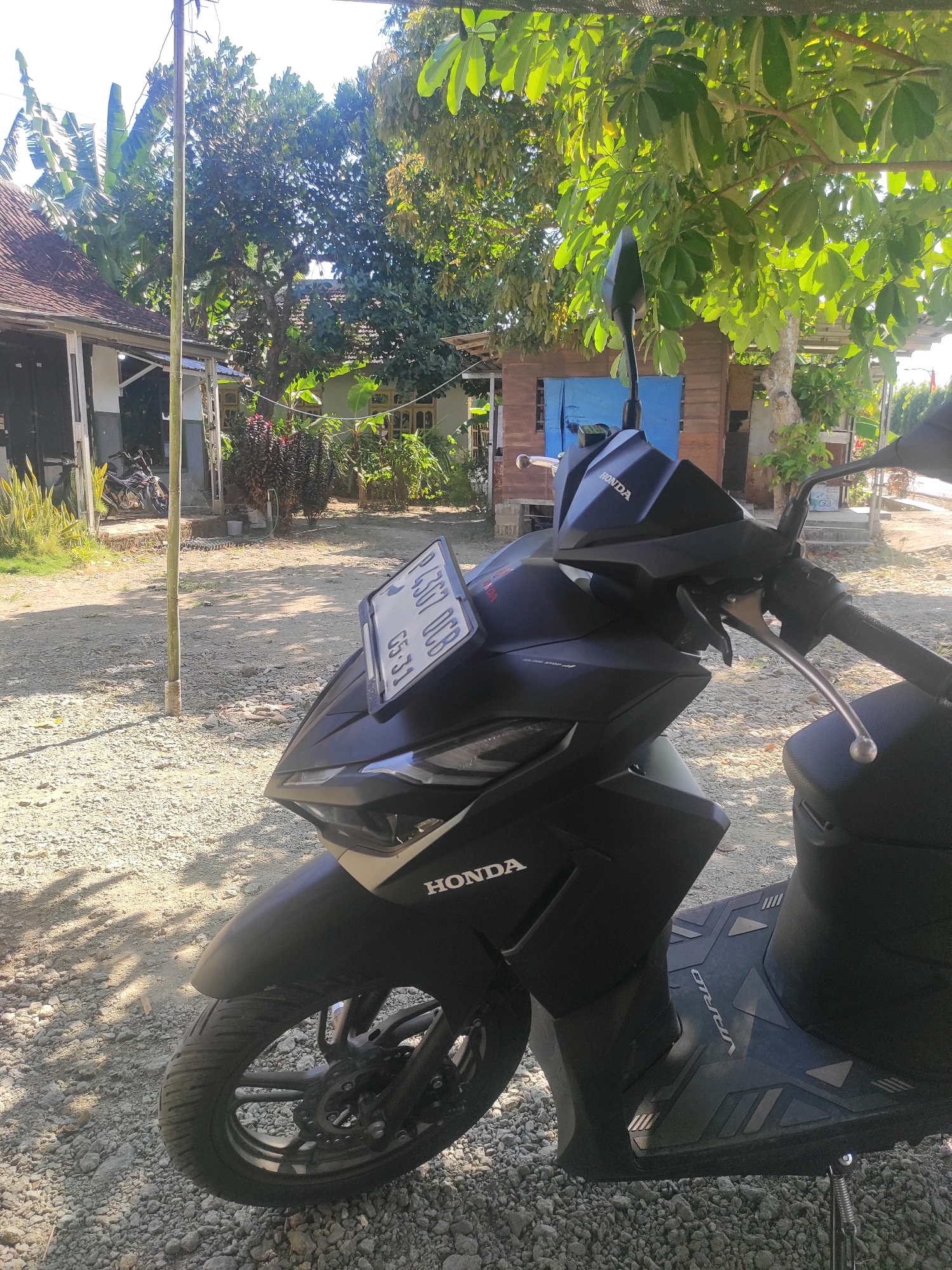 A black Honda scooter is parked on a gravel surface outside a residential building in Kandangan, Indonesia. The scooter is facing right, with its front wheel on the left side of the frame. The license plate, "P 4307 OCB," is visible on the front of the scooter. The scooter has a prominent "HONDA" logo on its front panel and "VARIO" branding on its floorboard. In the background, there are buildings with tiled roofs, trees with lush green leaves, and a wooden structure with a blue door. Sunlight casts shadows on the gravel.
