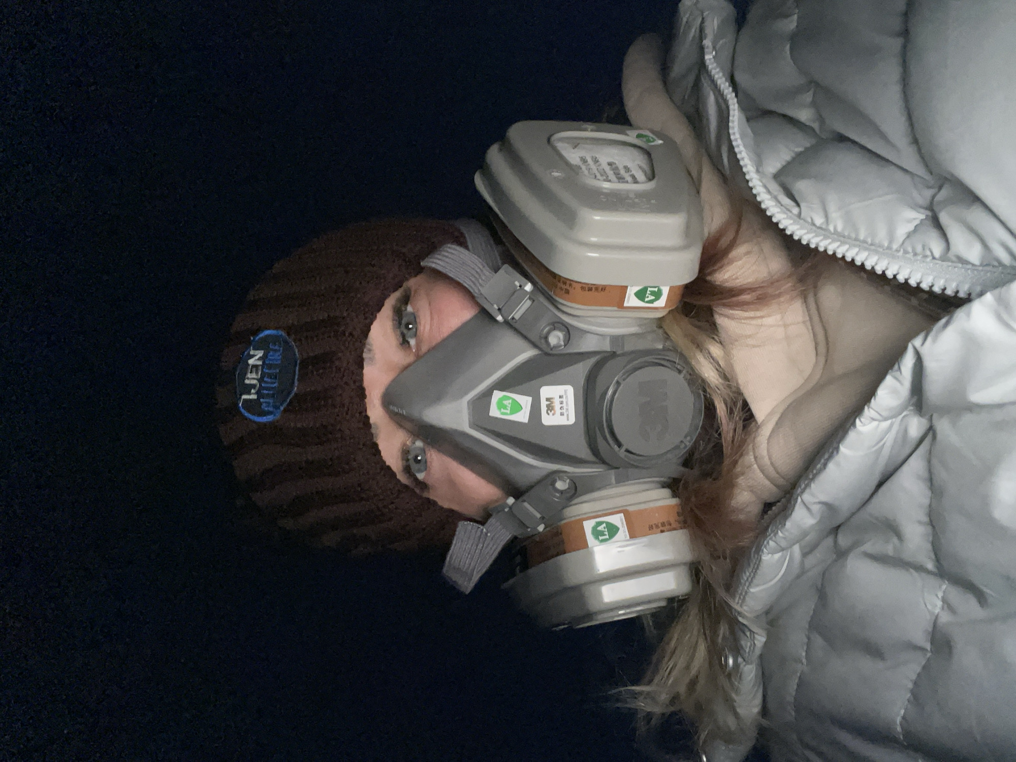 A person wearing a brown beanie, a gray respirator mask, and a silver puffer jacket is visible. The background is a dark blue. The beanie has a patch with blue writing on it. The mask is a gray 3M respirator. Part of the person's blonde hair is visible. The location is not discernible from the image.
