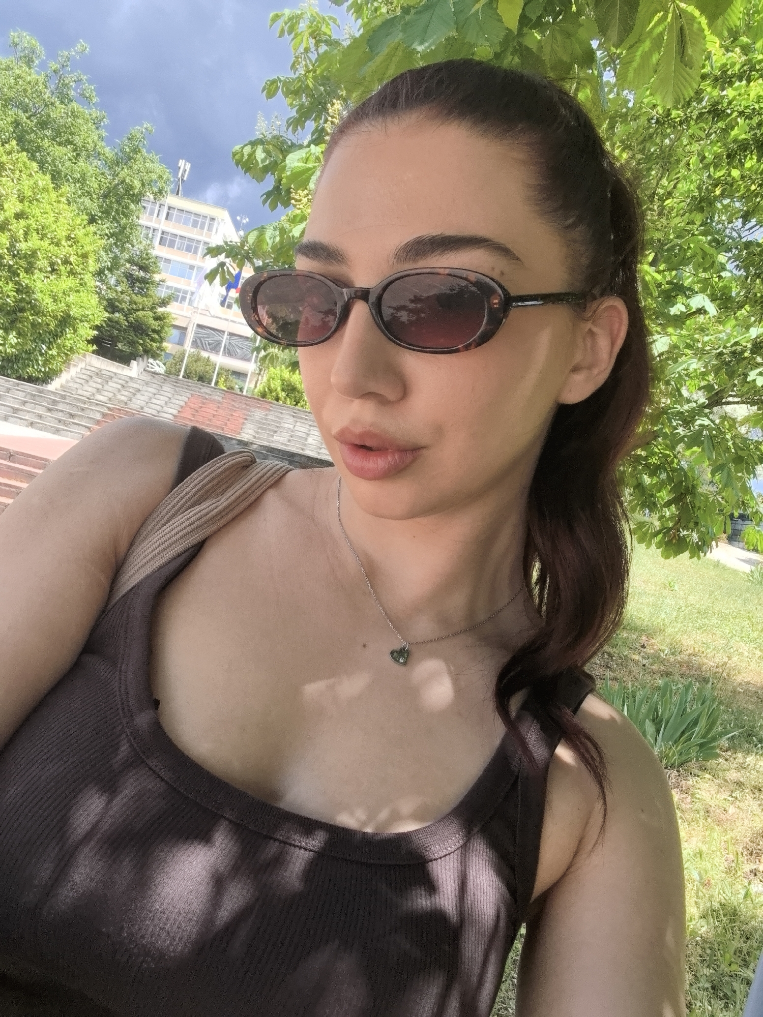 A woman is posing for a selfie. She has brown hair, pulled back into a ponytail with a few strands falling around her face. She wears oval-shaped sunglasses with dark frames and a brown tint. Her top is a dark brown, ribbed tank top. A silver necklace with a heart-shaped pendant is visible around her neck. The strap of a bag rests on her shoulder.
She is outside, under the shade of trees. A building can be seen in the background, with steps leading up to it. The location appears to be a park-like setting.