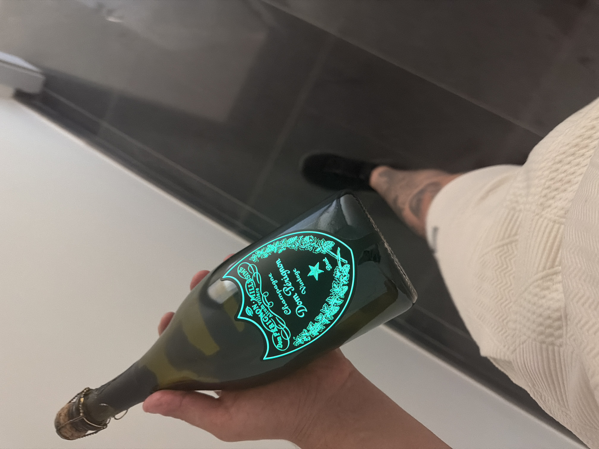 A person's hand holds a bottle of Dom Pérignon champagne, its label glowing with a cyan light. The hand and arm are visible, with tattoos on the forearm. The person is wearing white shorts. The background shows a modern interior with a dark gray tiled floor and a white wall. The scene is indoors, possibly in a house or apartment. There is no information about the location being in Dolno Ablanovo, Bulgaria.