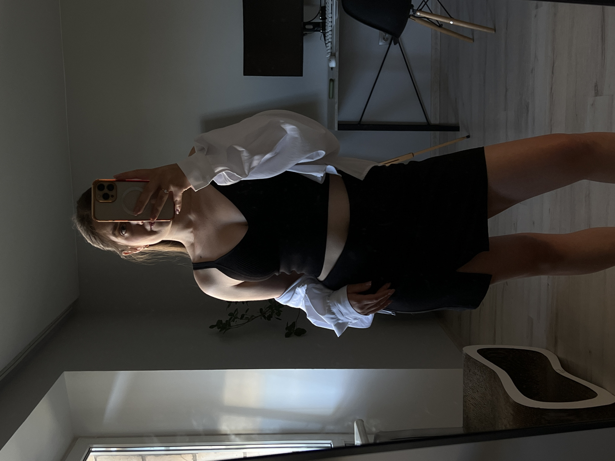 A young woman is taking a mirror selfie in an apartment. She is wearing a black ribbed tank top and a black skirt with a slit. A white unbuttoned shirt is draped over her shoulders. She is holding a smartphone to take the picture. In the background, there is a desk with a computer monitor, keyboard, and mouse. There is also a chair and a plant. The room is dimly lit, with light coming from a window on the left. The woman's reflection is visible in a large mirror on the left side of the image. The location is described as an apartment in the city of Ternopil, Ukraine.