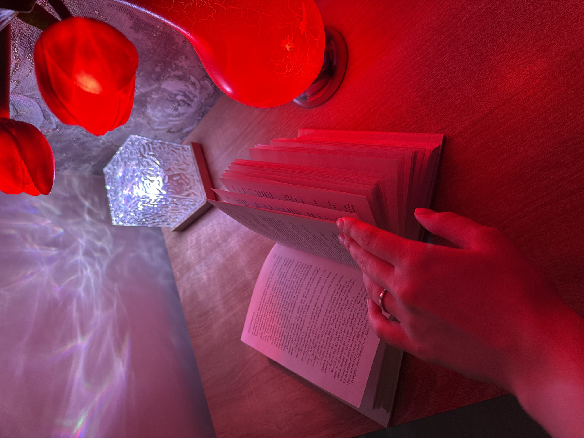 A person is reading a book under red lighting. The book is open and pages are being turned by a hand with a ring. Nearby are two light sources: a red lamp with a cracked glass effect and a cube-shaped lamp emitting a bright white light. The background appears to be a reddish-brown surface, possibly a table or desk, with faint patterns of light projected onto it.