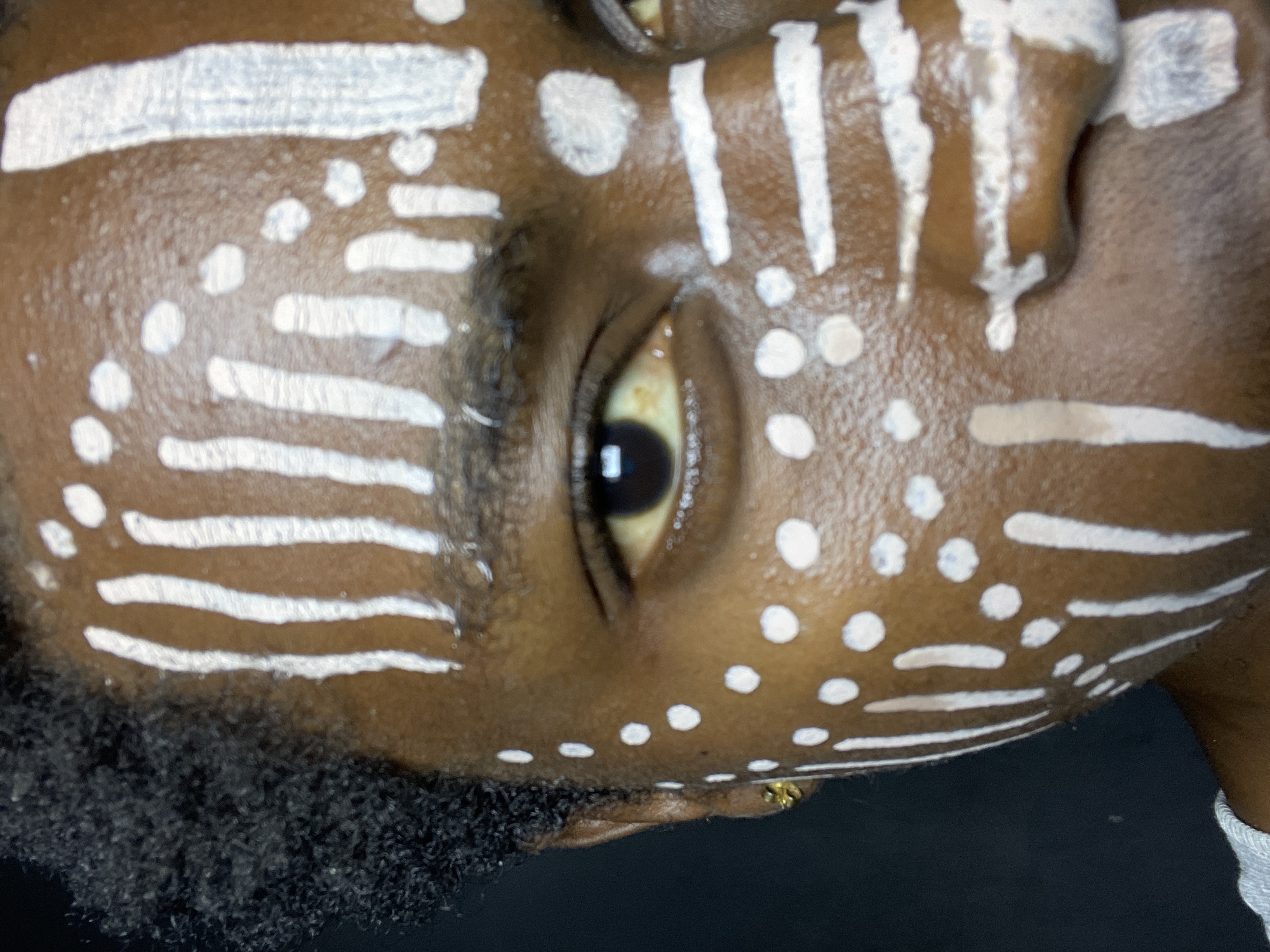 A close-up, tilted shot captures the side of a dark-skinned person's face, adorned with white paint. The design includes horizontal stripes and dots. Visible details include the person's eye, eyebrow, and a portion of their hair. An earring is visible. The background is dark. The location is unidentified; Nkpor, Nigeria is not indicated in the image.