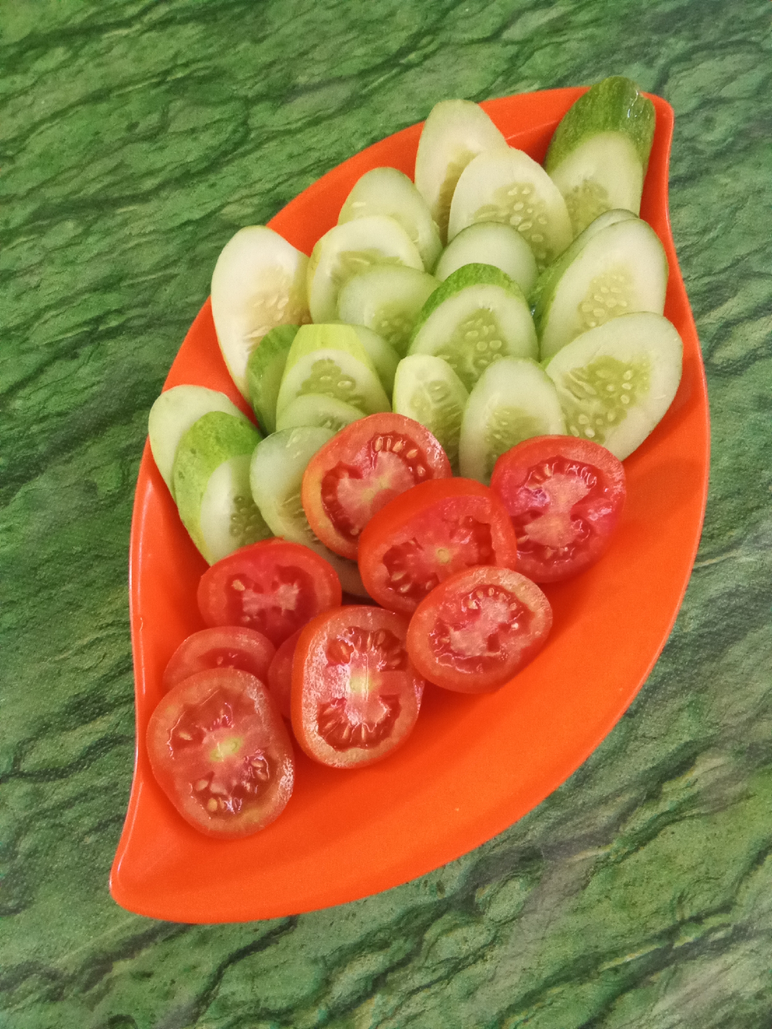 In a top-down, eye-level shot, a bright orange, leaf-shaped platter is filled with sliced vegetables. The platter rests on a textured, green surface that resembles marble. The platter is divided into two sections, with sliced cucumbers occupying the upper half and sliced tomatoes filling the lower half. The cucumbers are light green with visible seeds, while the tomatoes are a vibrant red. The platter and its contents are well-lit, with no shadows to obscure the details. The scene is clean and orderly, suggesting a prepared meal or appetizer. The location is identified as Subang, Indonesia, though no specific landmarks or environmental details beyond the platter and table are visible.