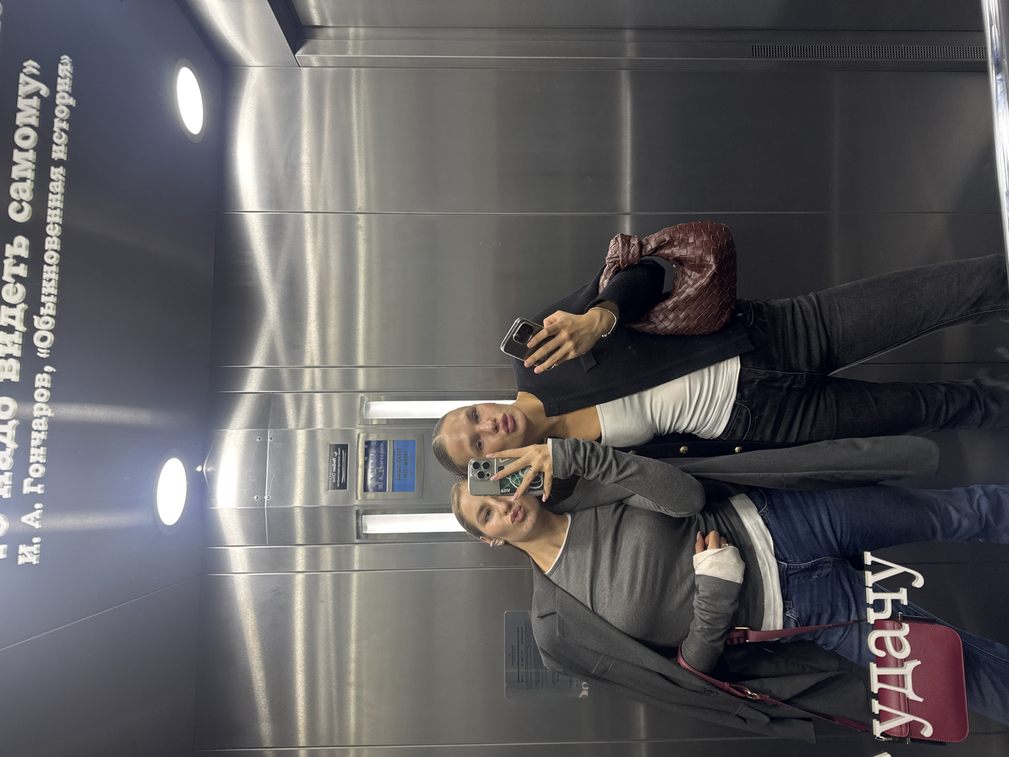 Two women are taking a mirror selfie in an elevator. The woman on the left is wearing a grey long-sleeved shirt, a grey blazer draped over her shoulders, and blue jeans. She has a red purse with a strap on her left side. The woman on the right is wearing a white tank top and black jeans. She has a brown woven bag on her right shoulder. Both women are puckering their lips. The elevator walls are made of brushed metal. There is text on the ceiling of the elevator.