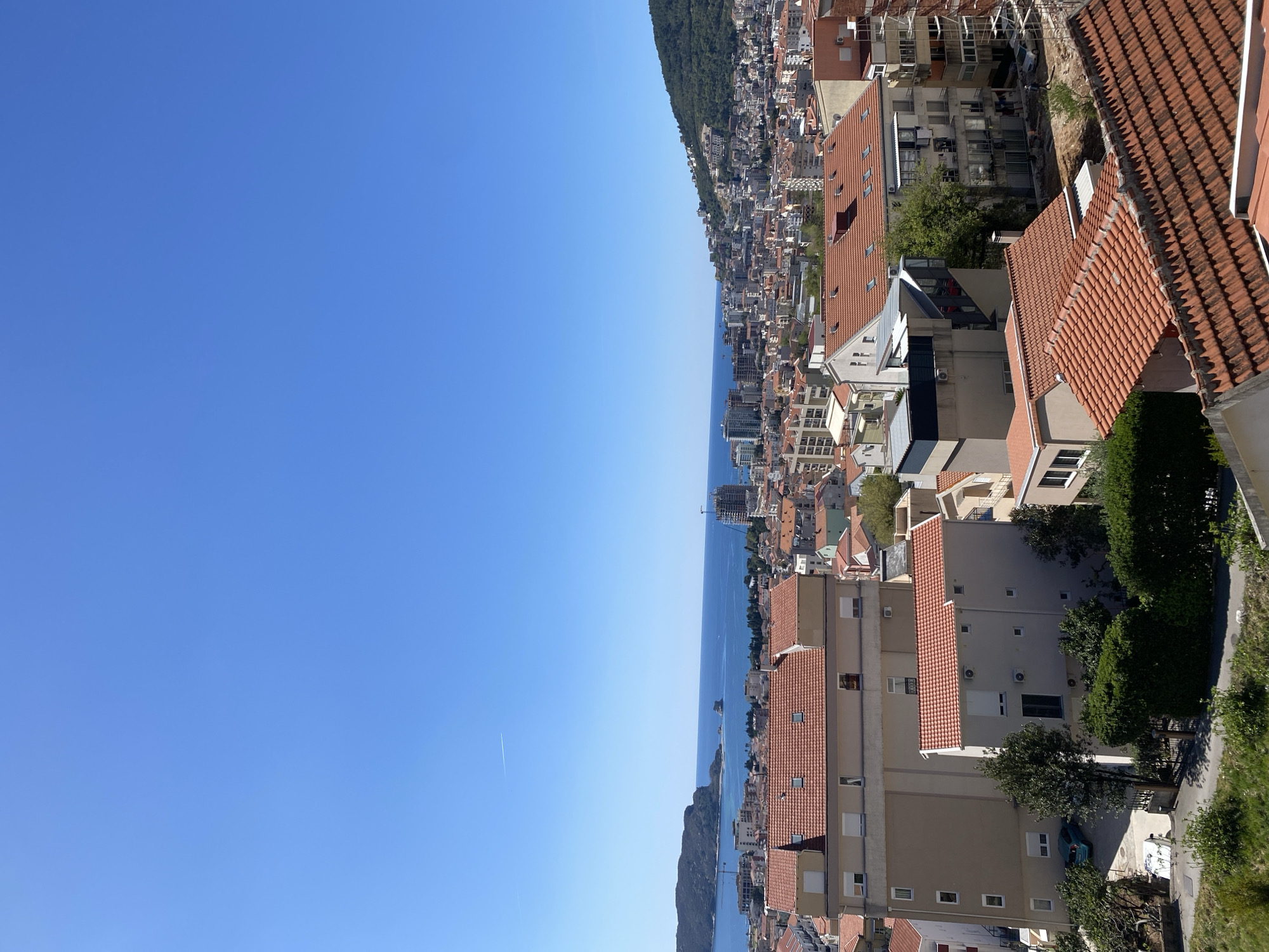 The image shows a coastal cityscape with a clear blue sky. A dense urban area with buildings of varying heights and red-tiled roofs stretches along the coastline. The Adriatic Sea is visible, separating the city from a green, mountainous area.
The location appears to be Budva, Montenegro, characterized by its coastal setting, red-tiled buildings, and mountainous backdrop.