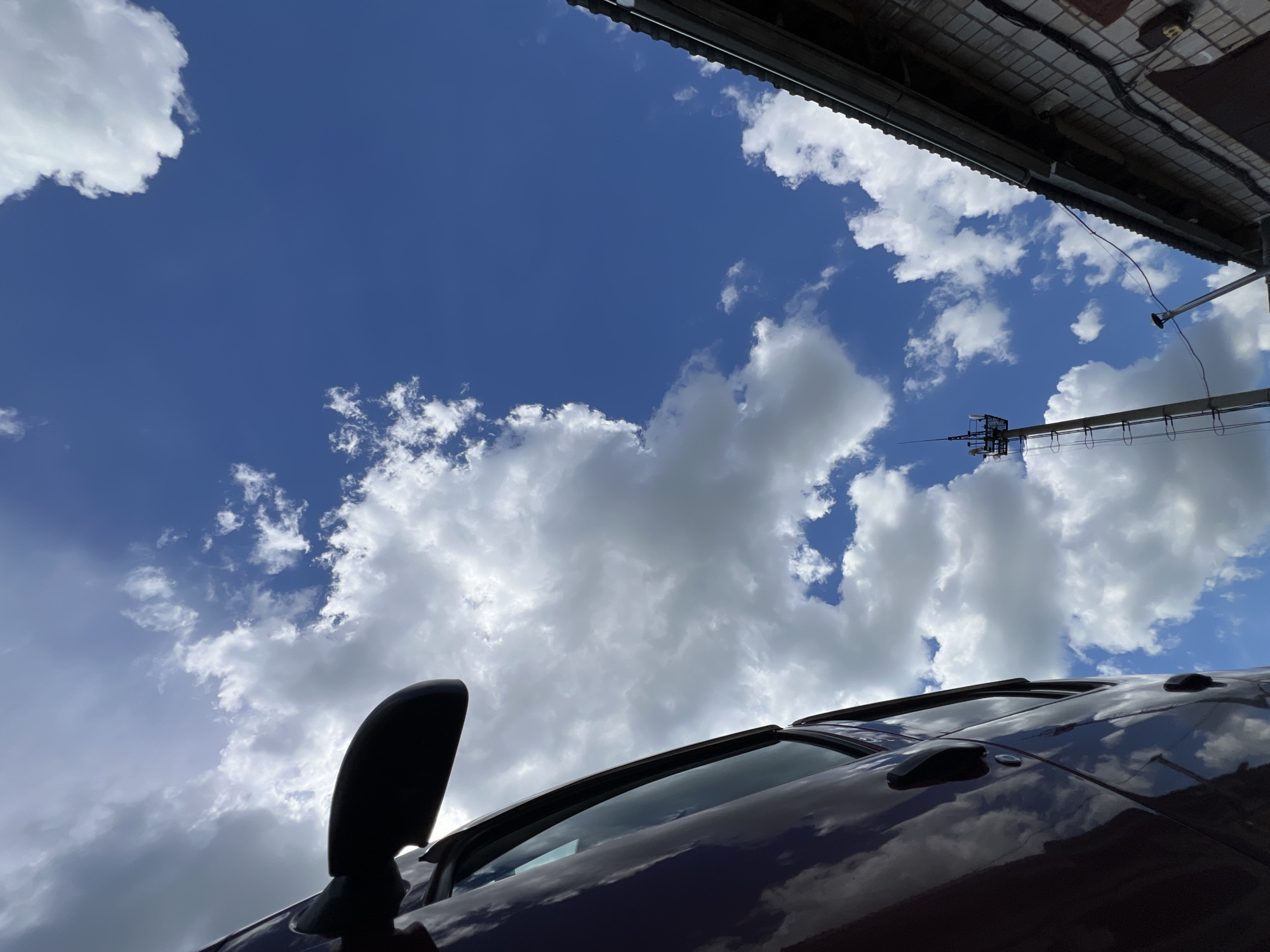The image depicts a low-angle view of a dark-colored car's roof and side mirror against a vibrant blue sky filled with fluffy white clouds. The car's glossy surface reflects the sky and clouds, creating a dynamic visual texture. In the upper portion of the frame, the underside of a dark, textured roof or overhang is visible, with some electrical wiring and a light fixture attached. A utility pole with equipment is also visible in the distance. The scene appears to be outdoors on a clear day, likely daytime, given the bright sky and clouds. The perspective suggests the photograph was taken from ground level, looking upwards. There are no people visible in the image, and no text is present. The setting is somewhat ambiguous but the presence of the utility pole and overhang might suggest an urban or semi-urban environment. The weather appears fair with scattered clouds.
