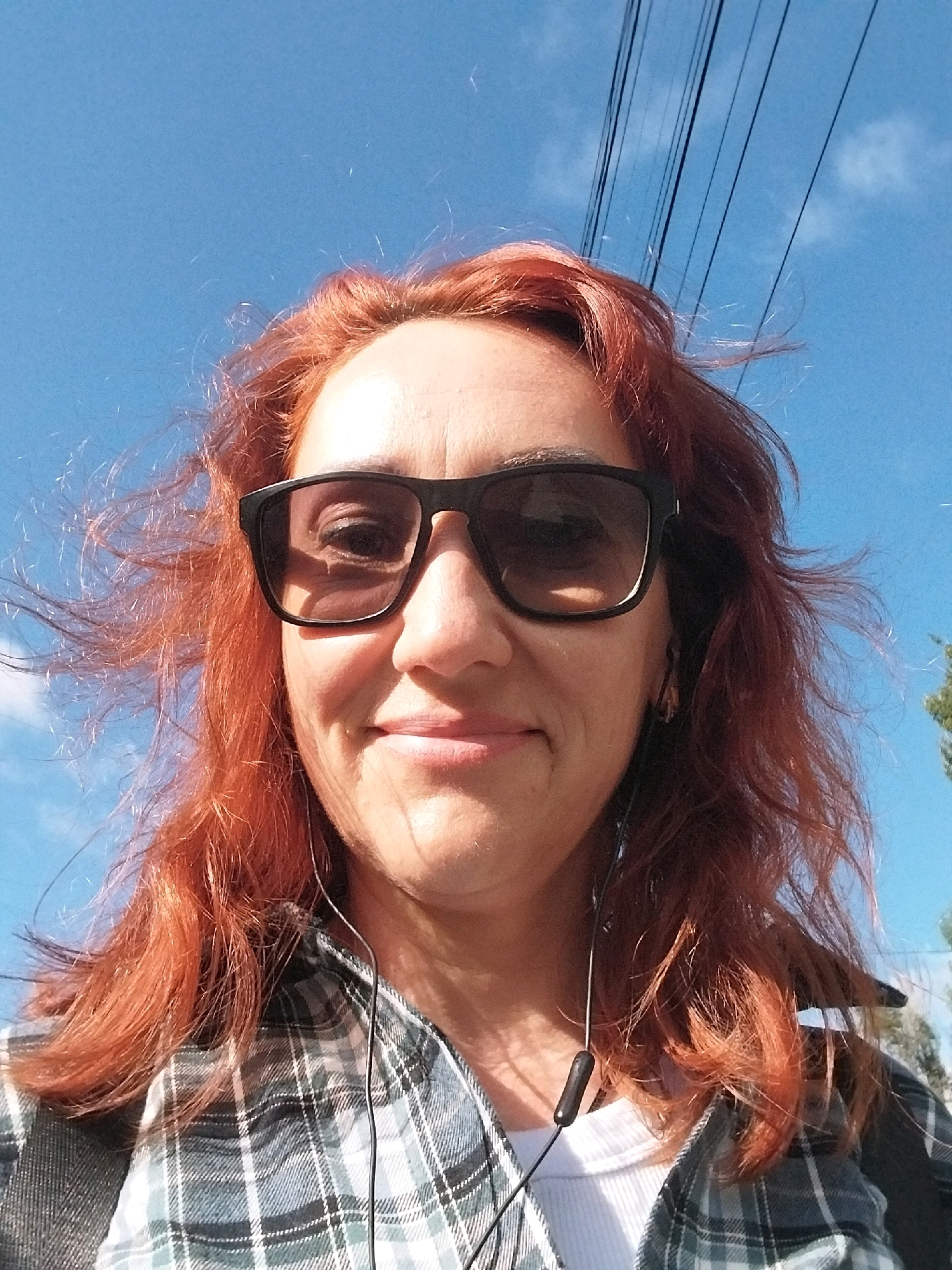 A medium shot selfie shows a woman with red hair and sunglasses looking at the camera outdoors. She is wearing a plaid shirt over a white shirt and has earbuds in. The background is a clear blue sky with some clouds and overhead power lines. The image is likely taken in Dnipro, Ukraine, based on the provided context.