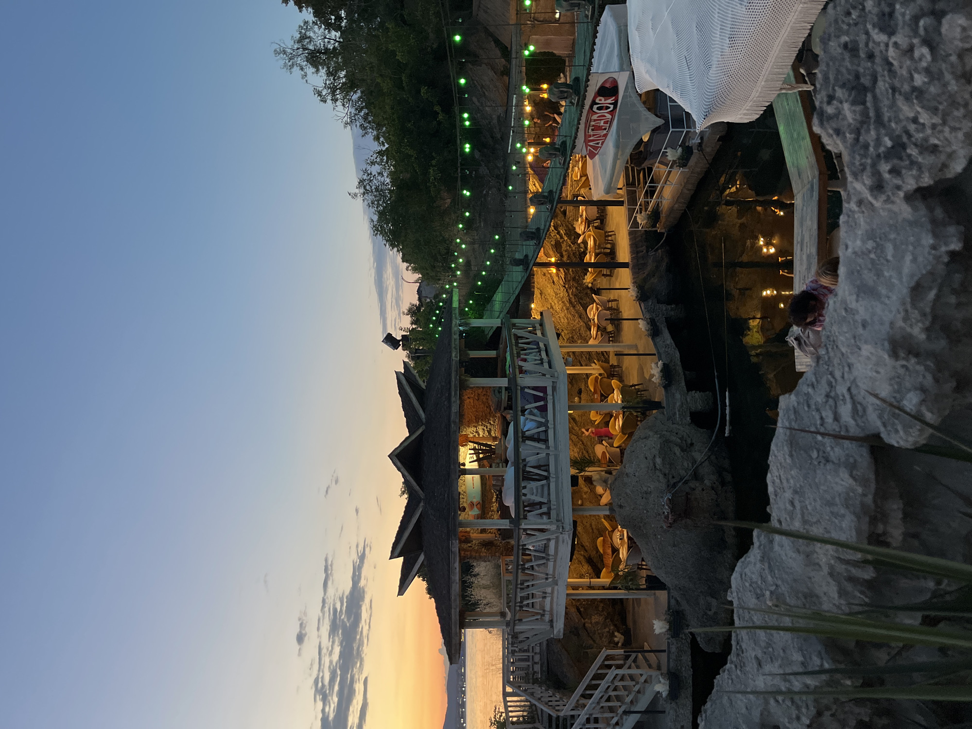 The image shows a scenic view of a seaside establishment in Kiten, Bulgaria, during dusk. A multi-level structure, likely a restaurant or resort, is built on a rocky coastline. The architecture features a central hexagonal building with a distinctive roof, surrounded by outdoor seating areas with tables and chairs. Wooden walkways and staircases connect different levels. Green string lights are visible along a suspended walkway, adding to the ambient lighting. In the foreground, large grey rocks and vegetation frame the scene. The sea is visible in the background, with a faint glow from the setting sun on the horizon. Several people are present, some seated at tables and others walking, indicating a lively atmosphere. The overall impression is one of a tranquil evening at a picturesque coastal location.