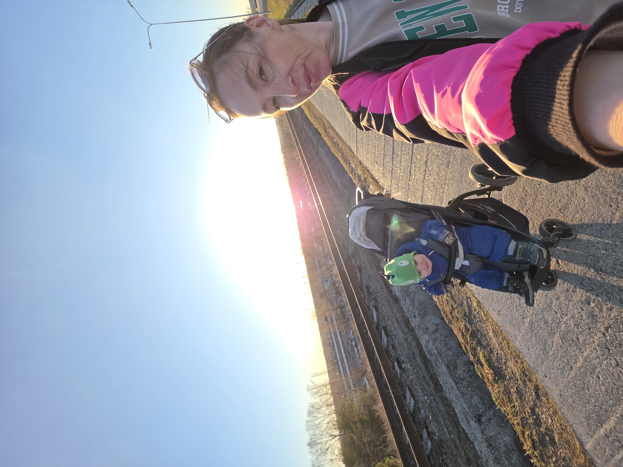 A woman is standing next to a railway track in Chaikovsky, Russia, pushing a child in a stroller. The child wears a green hat and a blue jumpsuit. The sky is clear, and the sun is setting, casting a bright light in the distance. The woman is wearing a pink and black jacket.