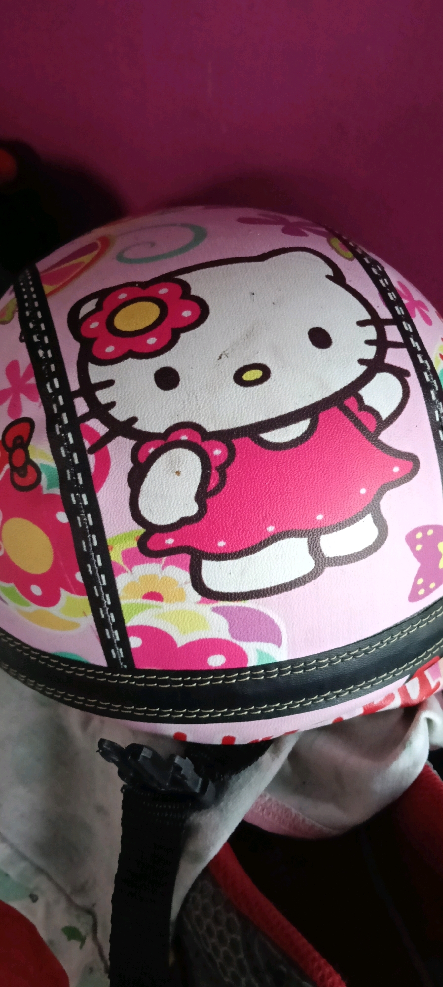 The image shows a close-up, eye-level view of a pink helmet adorned with a Hello Kitty character design. The helmet is decorated with flowers and bows in various shades of pink, yellow, and white. A black band with white stitching runs around the circumference of the helmet, and a black strap with a plastic buckle is visible in the foreground, attached to the helmet. The background is a solid pink color, suggesting an indoor setting or against a pink wall. The image does not provide any information about the location of Kuala Kapuas, Indonesia.