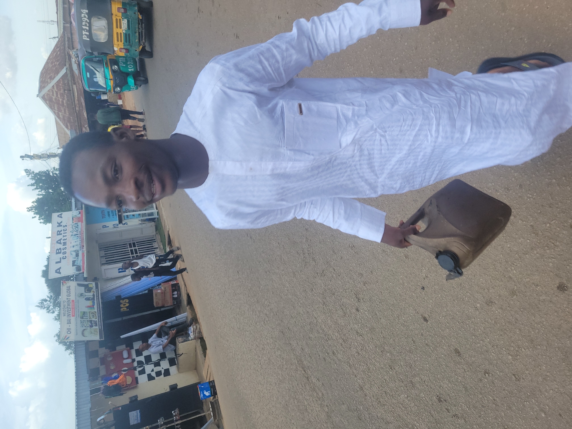 In Jos, Nigeria, a young man in a white kaftan walks along a paved street, carrying a dark brown jerrycan. The street is lined with shops, including one with signs for "CHI-BAI INVESTMENT GLOBAL" and "ALBARKA COSMETICS." A man sits outside one of the shops, and two young boys walk past. In the background, three auto-rickshaws are parked. The sky is overcast.