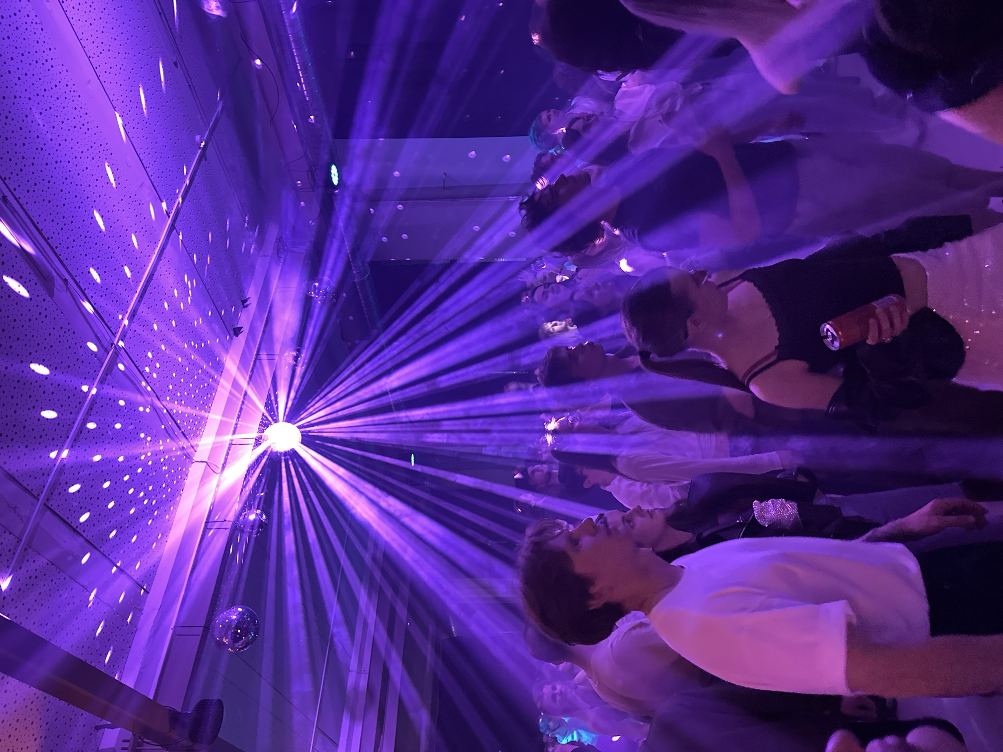 This image captures a crowded indoor social event, likely a party or club, under predominant purple lighting in the city of Minsk, Belarus. The venue features a textured ceiling, possibly acoustic tiles, with exposed ventilation ducts and structural elements. Multiple disco balls are suspended overhead; a prominent, brightly illuminated disco ball at the top-center casts numerous radial purple light beams across the scene, augmented by haze or smoke in the air. Smaller disco balls are also visible to the left.
The scene is filled with numerous young adults, mostly standing and socializing. In the foreground to the left, a young man with short dark hair and a gold neck chain wears a white t-shirt and looks towards the right, slightly upwards. In the foreground to the right, a young woman with dark hair tied in a ponytail, wearing a black spaghetti strap top and a light-colored, possibly sequined, skirt, holds a red beverage can in her left hand and looks down. Her left arm displays a tattoo.
Mid-ground individuals include a person in a dark t-shirt and light pants to the left of the woman with the can, and another individual with blue hair further back on the right. People are dressed in casual attire, consistent with a contemporary social gathering. The collective posture and arrangement of the individuals suggest an active, social environment. A light fixture emitting a green-blue light is visible on a wall in the mid-ground right. No overt interactions are discernible, but the proximity of individuals indicates a group gathering.