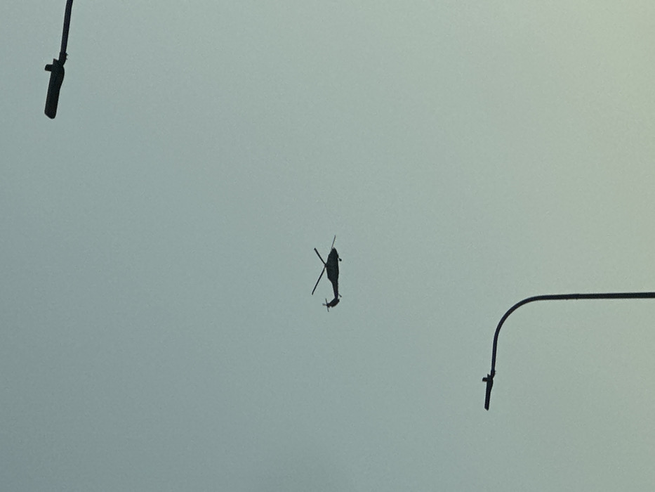 The image displays a utility helicopter in flight, silhouetted in black against a uniformly light blue, clear sky. The helicopter, which appears to be a Black Hawk variant, is positioned slightly to the left of the image's horizontal center, with its main and tail rotors distinctly visible. In the foreground, two black, curved street light poles are partially visible as silhouettes: one enters the frame from the bottom-left edge, curving upwards, and the other from the top-right edge, curving downwards. No individuals or specific ground features are discernible in the frame. The scene is located within the Galil Elyon Regional Council, Israel.