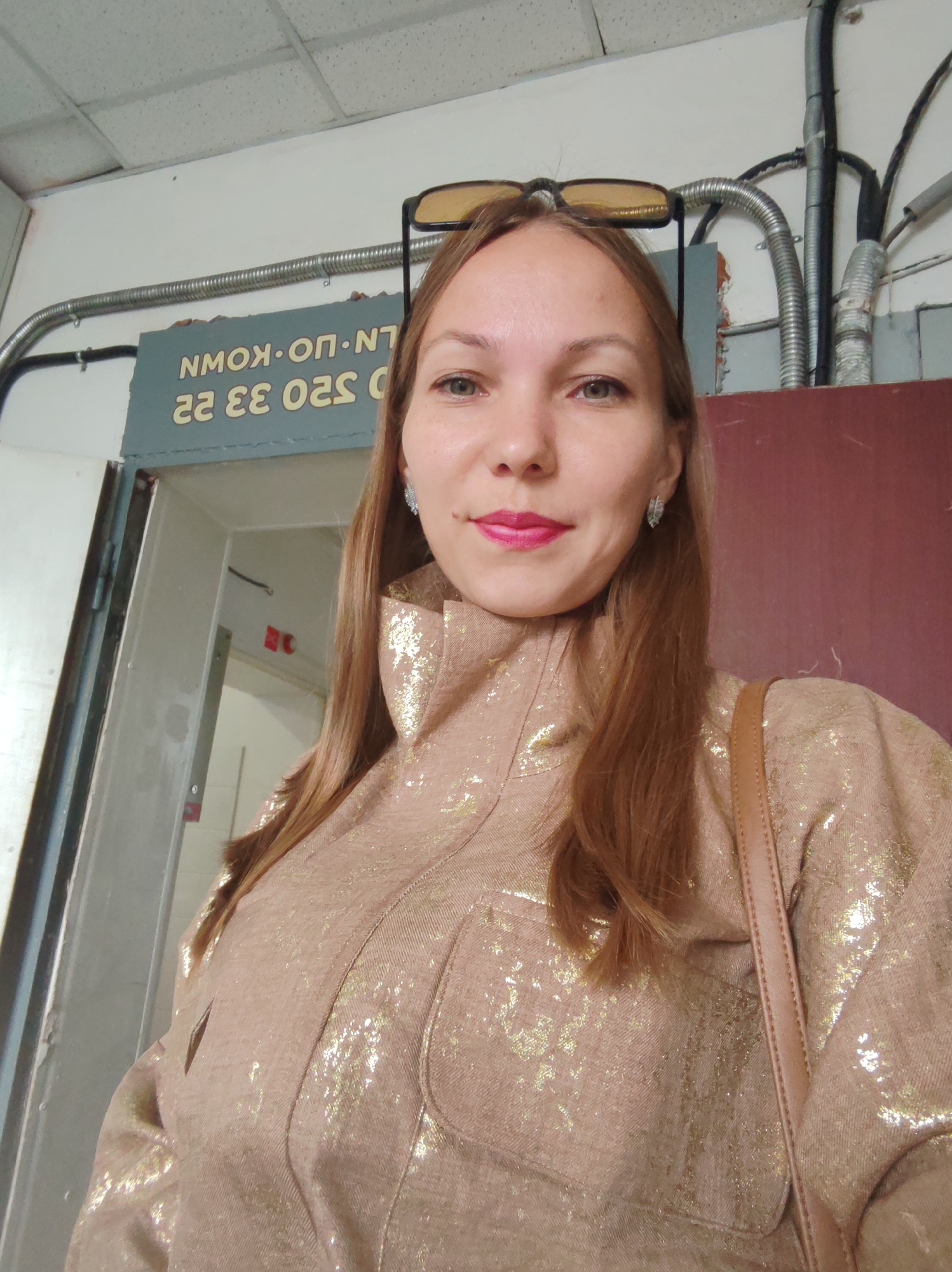 The image shows a close-up selfie of a woman with long, brown hair. She is wearing a tan jacket with a gold speckled pattern, and a brown purse strap is visible over her shoulder. She has on bright pink lipstick and silver earrings. In the background, there is a sign with reversed Cyrillic text that reads "NMOK.ON.NT" and "22 33 OS2 0". Above the sign, conduit pipes are mounted on the wall. The woman is standing in an indoor environment, possibly a hallway or corridor, with a doorway visible behind her, leading to a dimly lit room. The overall scene is casual and personal, suggesting the photo was taken by the subject herself. There is no discernible information in the image that would indicate a specific location within Syktyvkar, Russia.