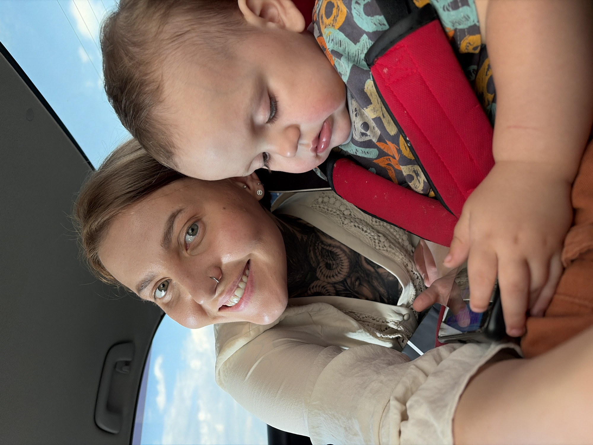 A medium shot shows a woman and a baby in a car. The woman is in the foreground, looking at the camera with a smile. She has blonde hair, green eyes, and a nose ring. She is wearing a cream-colored shirt with lace detailing and has tattoos on her neck. To her right, the baby is in a car seat, looking down at a smartphone held by the woman's hands. The baby has light brown hair and is wearing a colorful shirt. The car's interior is visible, with the sky and clouds in the background through the windows. The scene appears to be in Chaikovsky, Russia, based on the provided context.