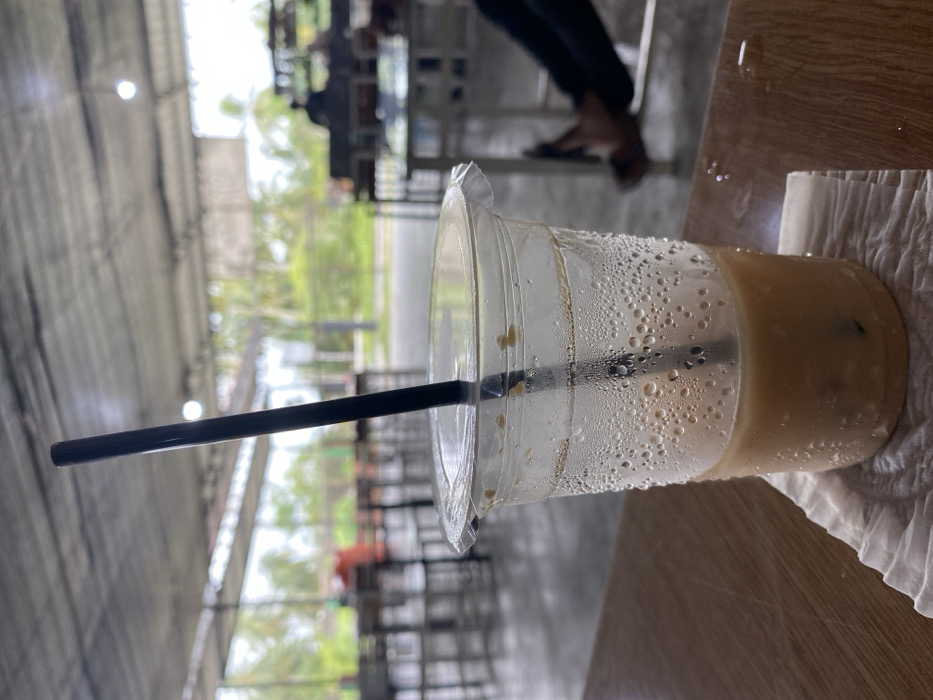 A partially consumed iced beverage in a clear plastic cup, containing a black straw and ice cubes, rests on a white paper napkin on a light brown table. Condensation is visible on the cup's exterior, and water droplets are present on the table surface. The scene is located within a semi-open commercial establishment in Aluebutuha, Indonesia, featuring a grey corrugated roof supported by metal beams and a concrete floor. Multiple light-colored tables and chairs are arranged throughout the space. In the middle-ground, the lower legs and feet of an individual, wearing dark pants and sandals, are visible to the right. A blurred figure is seated at a table in the background. Beyond the structure, a road runs parallel to lush tropical vegetation, including palm trees.