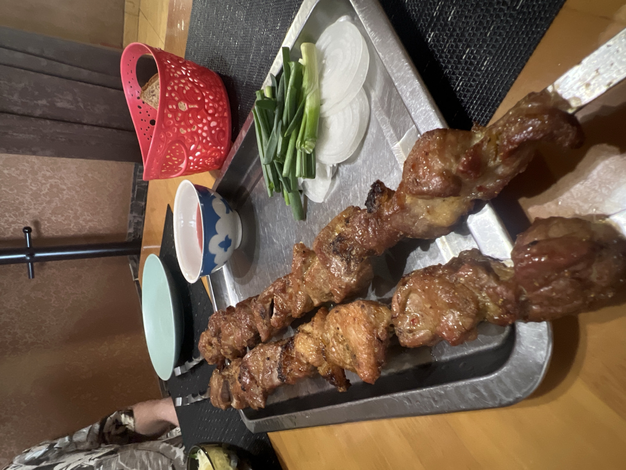 The image shows a meal served on a metal tray. There are two skewers of grilled meat, cut green onions, and sliced white onions on the tray. A red plastic bread basket, a small bowl with a blue and white design, and a light green plate are also visible. The tray sits on a surface that appears to be a table. The exact location and time are not discernible.