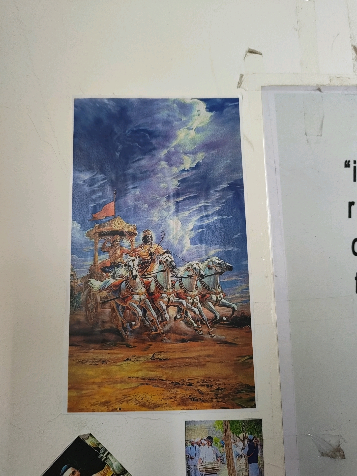A framed print, depicting a scene from the Bhagavad Gita, is affixed to a white, textured wall. The print features a chariot pulled by four white horses, with two figures in traditional Indian attire visible within the chariot. The sky in the print is dramatic, with swirling clouds in shades of blue, purple, and yellow, illuminated by sunlight. The ground below is rendered in earthy tones of brown and orange. To the right of the print, a white piece of paper with text in black is partially visible. Below the main print, a smaller, diagonal print shows a group of men in traditional clothing, one of whom is playing a drum. The overall scene appears to be an interior wall in a residence or public building. This image was captured in Tathawade, India.