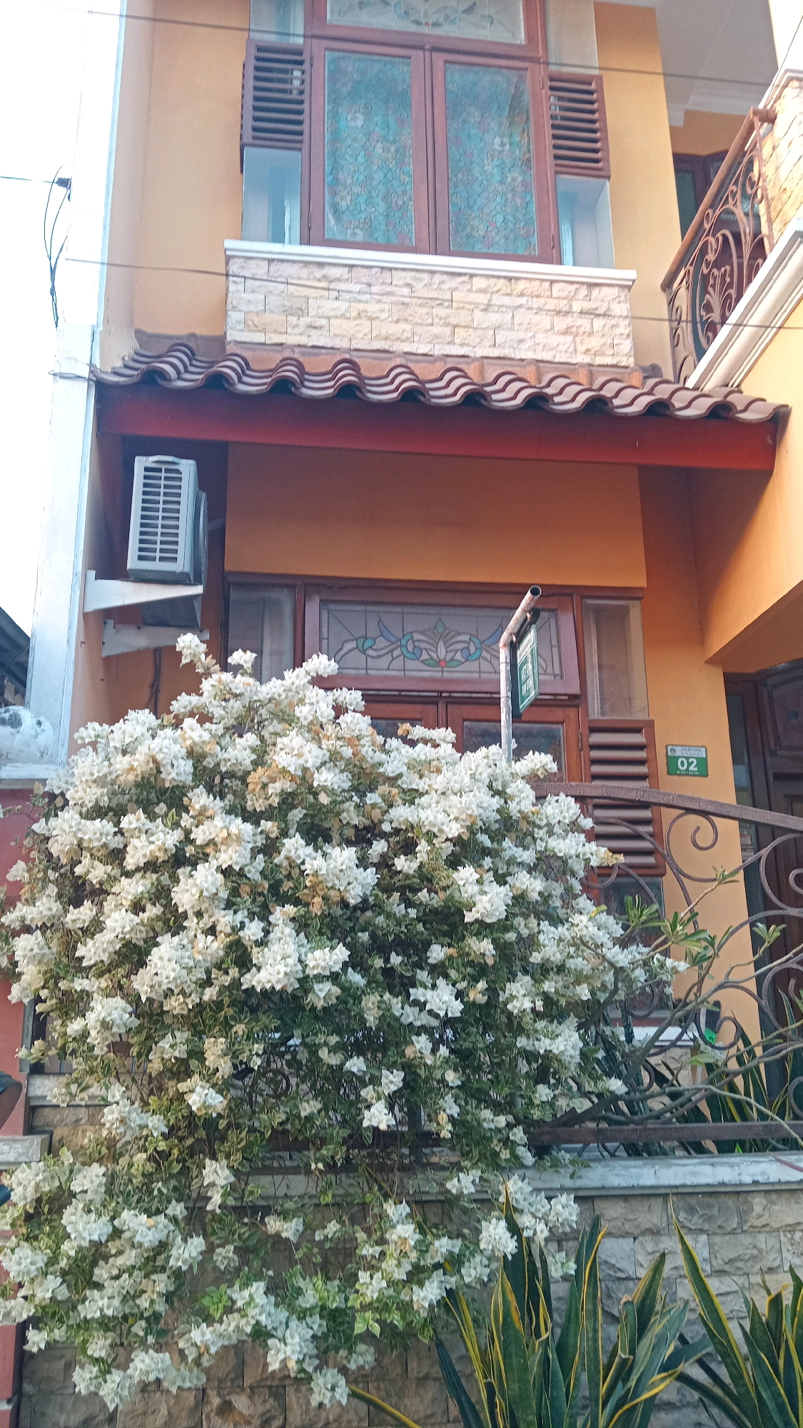 The image shows the facade of a building in Manyarsidomukti, Indonesia, with a large bush of white bougainvillea flowers in the foreground. The building has a yellow-orange exterior, with dark brown wooden window frames and doors. Above the entrance, there is a decorative stained-glass window. An air conditioning unit is visible mounted on the wall to the left of the entrance. A metal fence with ornate scrollwork separates the building from the foreground. The building is two stories high, with the upper floor featuring a dark brown window with louvered shutters and decorative stained glass. A tiled roof with a curved design is visible between the two floors. A green sign with white text and the number "02" is attached to a metal post near the entrance. In the lower right corner, the leaves of a snake plant are visible. The overall scene depicts a residential or commercial building entrance adorned with flowering plants and architectural details.