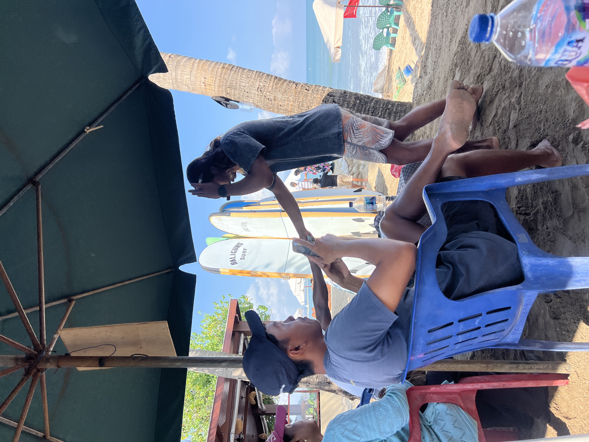 This image depicts a beach scene in Kuta, Indonesia, under a large green umbrella providing shade. In the foreground, two young men are seated on plastic chairs, facing away from the camera. The man on the left wears a blue t-shirt and a dark baseball cap, and is holding a smartphone. The man on the right wears a dark t-shirt and patterned shorts and appears to be interacting with the man on the left, possibly showing him something on the phone. Their legs and feet are visible, resting on the sandy beach.
Behind them, a line of surfboards with "BALIGUNS SURF" branding stands upright. A tall palm tree is visible to the right of the surfboards. In the background, the ocean is calm, with a few beach chairs and umbrellas on the sand. The sky is clear and blue. The overall atmosphere suggests a casual beach setting, possibly a surf school or rental spot. The presence of surfboards and the beach environment clearly places the location in Kuta, a well-known surfing destination.
