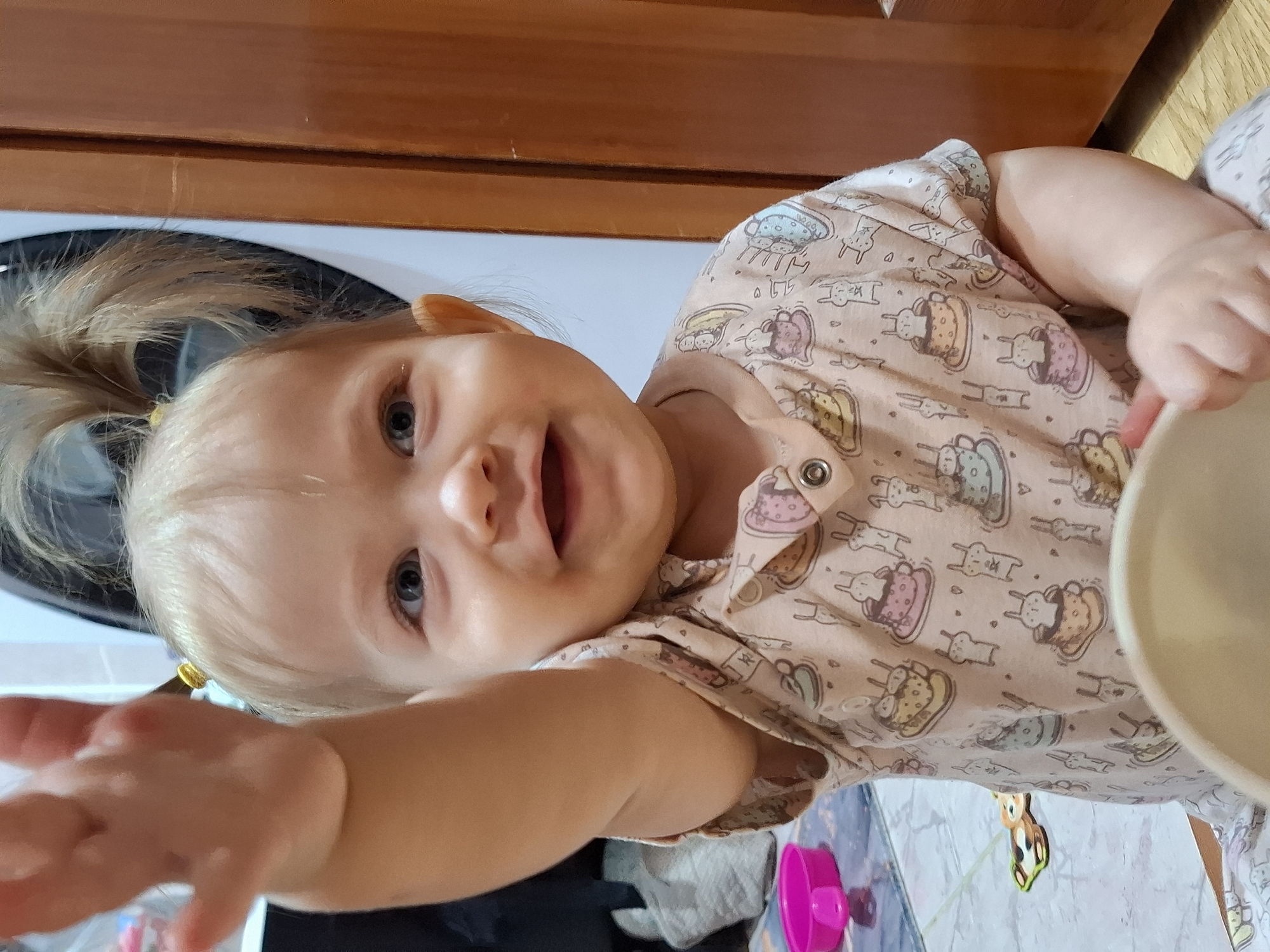 A close-up, slightly tilted portrait captures a joyful baby indoors. The infant, with fair skin, light blonde hair pulled into a ponytail, and bright blue eyes, is smiling widely with an open mouth, conveying happiness and engagement.
The baby is dressed in a light-colored, possibly peach or beige, onesie or top featuring a repeating pattern of whimsical cartoon animals, such as bunnies and other creatures, some appearing within cups or bowls. The baby's left hand (right side of the image) is positioned near a white bowl, while the right arm (left side of the image) is playfully outstretched towards the viewer, fingers spread, as if reaching out.
The background reveals elements of a home environment. Directly behind the baby's head is a dark circular opening, indicative of a washing machine's door seal. Above this, and to the right, warm-toned wooden cabinetry or furniture is visible. Below the baby's arms, there is a light-colored, possibly patterned, surface, alongside a bright pink cup or toy and a small, colorful animal-shaped toy.
The scene depicts a candid, cheerful moment, likely during daytime given the natural light. The overall impression is one of a happy, playful baby interacting with the camera in a domestic setting in Chaikovsky, Russia. No visible text is present.