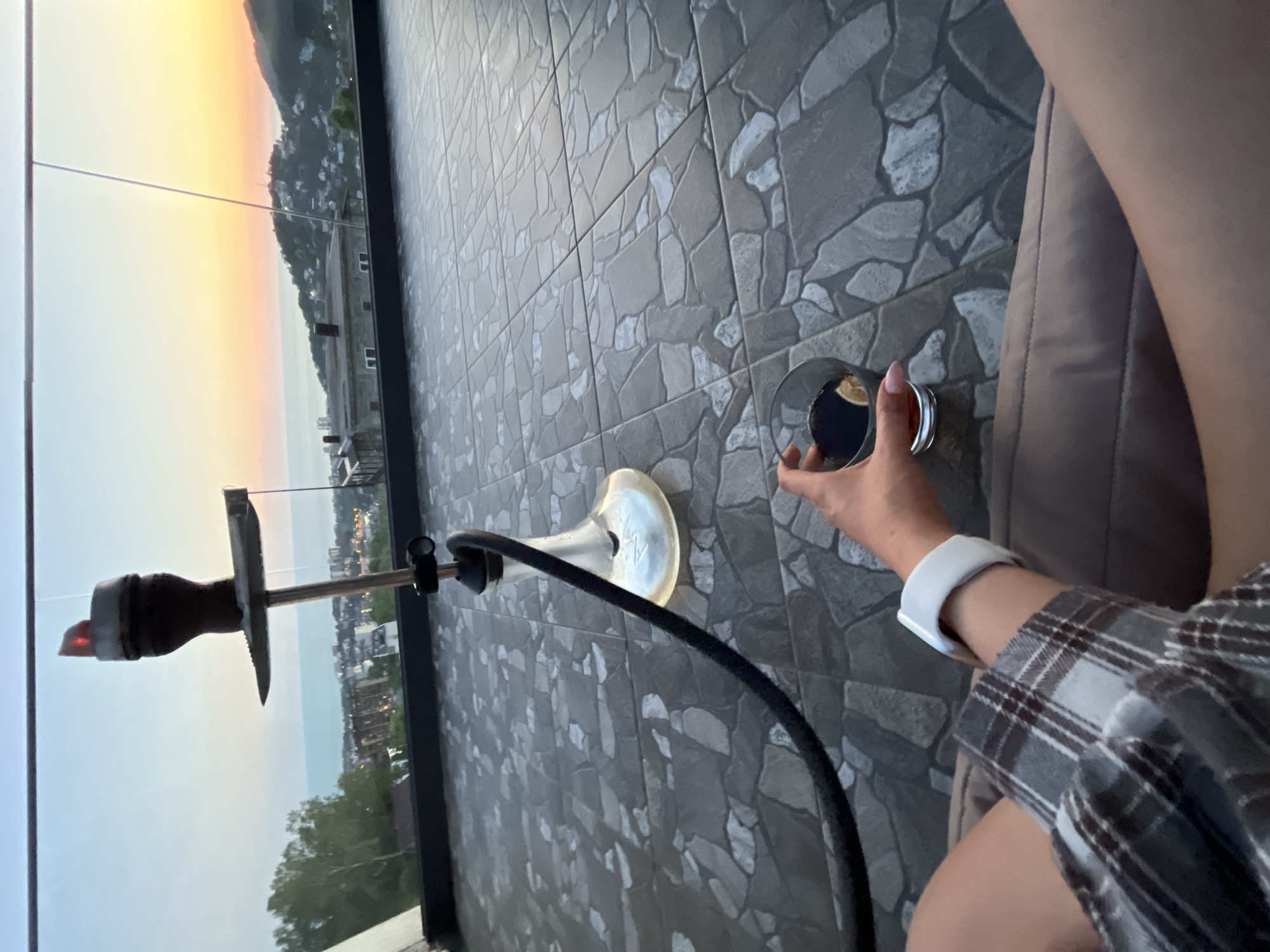 The image depicts a first-person perspective from an elevated outdoor terrace or balcony at dusk, presenting a panoramic view of a coastal city. In the immediate foreground, a person's bare legs, clad in shorts, are visible as they are seated, likely on a lounge chair. The person's right arm, wearing a white smartwatch on the wrist and a grey plaid long-sleeved shirt, holds a clear glass containing a dark liquid with a slice of lime or lemon. The flooring is composed of square grey tiles with irregular, light grey patterns.
A modern hookah with a transparent, bell-shaped glass base and a black hose rests on the tiled floor. Its dark ceramic bowl, positioned at the top of a metal stem, contains visible red, glowing coals. Beyond the hookah, a clear glass railing defines the edge of the terrace, overlooking a sprawling urban landscape.
The background reveals a city with numerous buildings, some of which display illuminated windows or streetlights, indicating evening. This city is situated along a coastline, with the sea extending to the horizon under a twilight sky. The sky transitions from a soft blue overhead to vibrant orange and pink hues near the horizon, characteristic of a sunset. Green, forested hills and mountains rise behind the urban area, blending into the colorful sky. The overall scene suggests a relaxing evening setting in a coastal mountain region, consistent with the geography near Baranovka, Russia, which is situated in the Greater Sochi area and known for its mountainous terrain and proximity to the Black Sea.