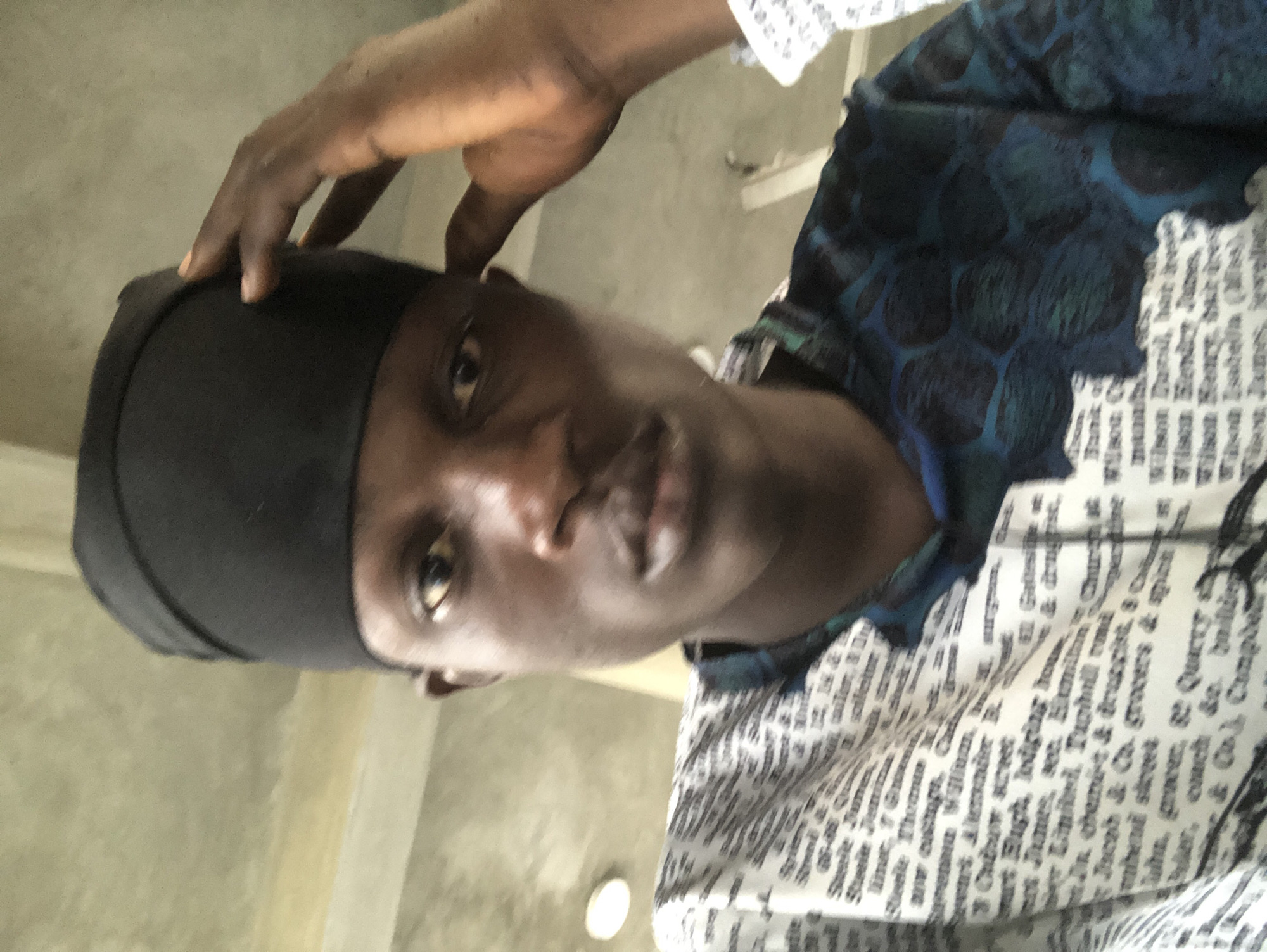 A dark-skinned male with a black cap is captured in a close-up, selfie-style photograph. He is wearing a white shirt with black text and a blue and green patterned shirt underneath. His right hand is resting on the top of his head, near the cap. The background appears to be an indoor setting with a concrete ceiling and a light fixture visible on the left. The image quality is slightly blurry, but the subject's facial features are discernable, including his eyes and lips. The overall lighting suggests an indoor environment with artificial light. The location is identified as Ado, Nigeria.