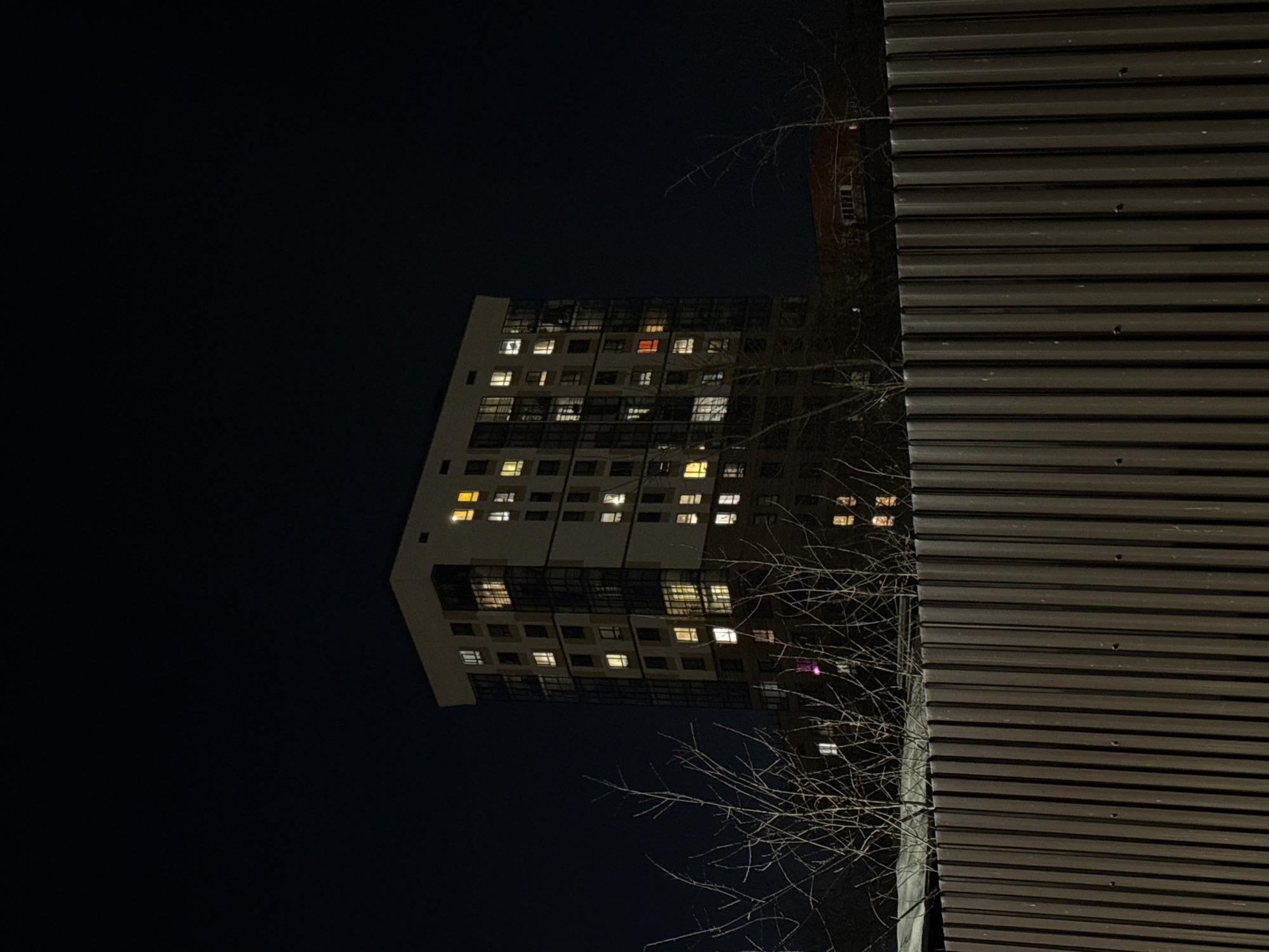 The image captures a nighttime urban scene in Izhevsk, Russia, likely photographed in autumn or winter given the bare tree branches. A high-rise apartment building with illuminated windows is prominently featured, contrasting with the dark sky. Adjacent to the apartment building is a metal-clad structure, possibly a fence or wall, characterized by horizontal ribbed panels. The lighting is primarily from the building's windows, with some subtle light reflecting off the metal structure. No people are visible in the scene.