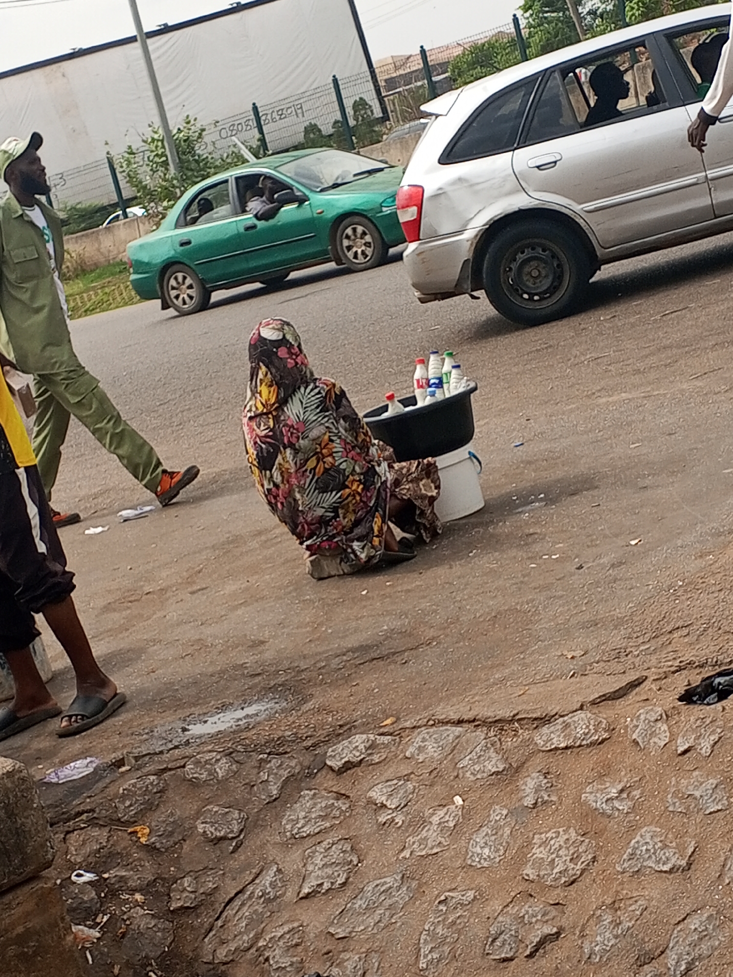 A street scene in what is likely Abuja, Nigeria, based on cultural context, features a woman squatting on the side of a paved road, displaying a bucket containing several bottles of liquid, suggesting she is a vendor selling drinks. She is dressed in a patterned, floral-print robe with a matching head covering. Two men are visible passing by her; one is wearing a green outfit and the other has black pants with a yellow stripe down the side. The road is flanked by moving vehicles, including a green sedan and a silver hatchback. The road surface is cracked, with some loose debris and trash present. The backdrop includes a building, a fence, and scattered vegetation. A concrete section with cobblestone patterns is present in the foreground.