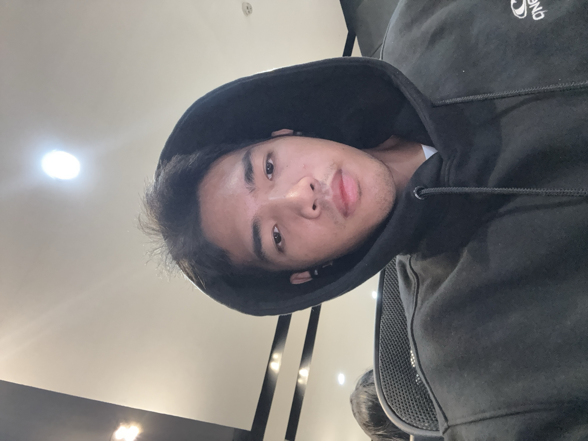 The image is a selfie taken indoors, likely in an office environment, of a young Southeast Asian male. He is wearing a black hooded sweatshirt with a white t-shirt underneath, and a hood is pulled up over his head. He is also wearing an earbud in his right ear. The background shows ceiling lights and what appears to be office furniture, including the back of an office chair with a mesh headrest. There is another person's head visible behind the subject to the left, also appearing to be in an office chair. The lighting suggests overhead fluorescent lights. The subject is looking directly at the camera. There is no specific visual information that can definitively place the location within Davao City, Philippines.