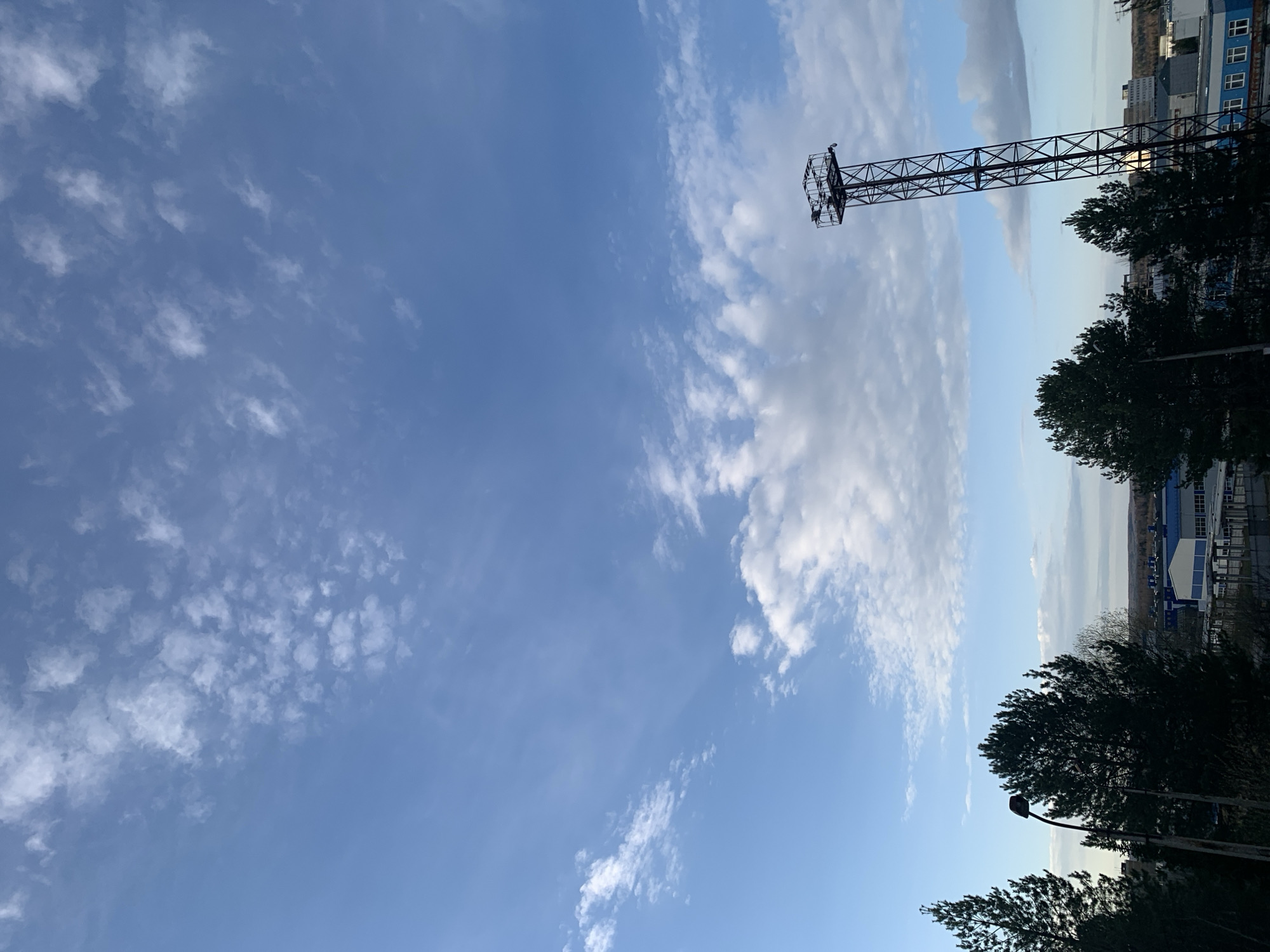 The image captures an expansive view of a blue sky, adorned with scattered white clouds of various formations, ranging from wispy cirrus-like clouds to more defined cumulus types. The lighting suggests it is daytime, possibly late afternoon or early morning.
On the right side of the frame, a tall, black, lattice-like metal structure, possibly a billboard frame or an industrial tower, extends vertically, featuring some equipment or lights at its uppermost point. Below and to the right of this structure, the dark silhouettes of trees are visible, with a street light pole partially discernible among them.
In the background, beyond the trees and the prominent metal structure, a collection of industrial-looking buildings with blue and grey exteriors can be seen, indicating an urban or industrial setting, consistent with the location context of Chaikovsky, Russia. One building has a distinct row of small windows. No human activity or visible text is present in the image.