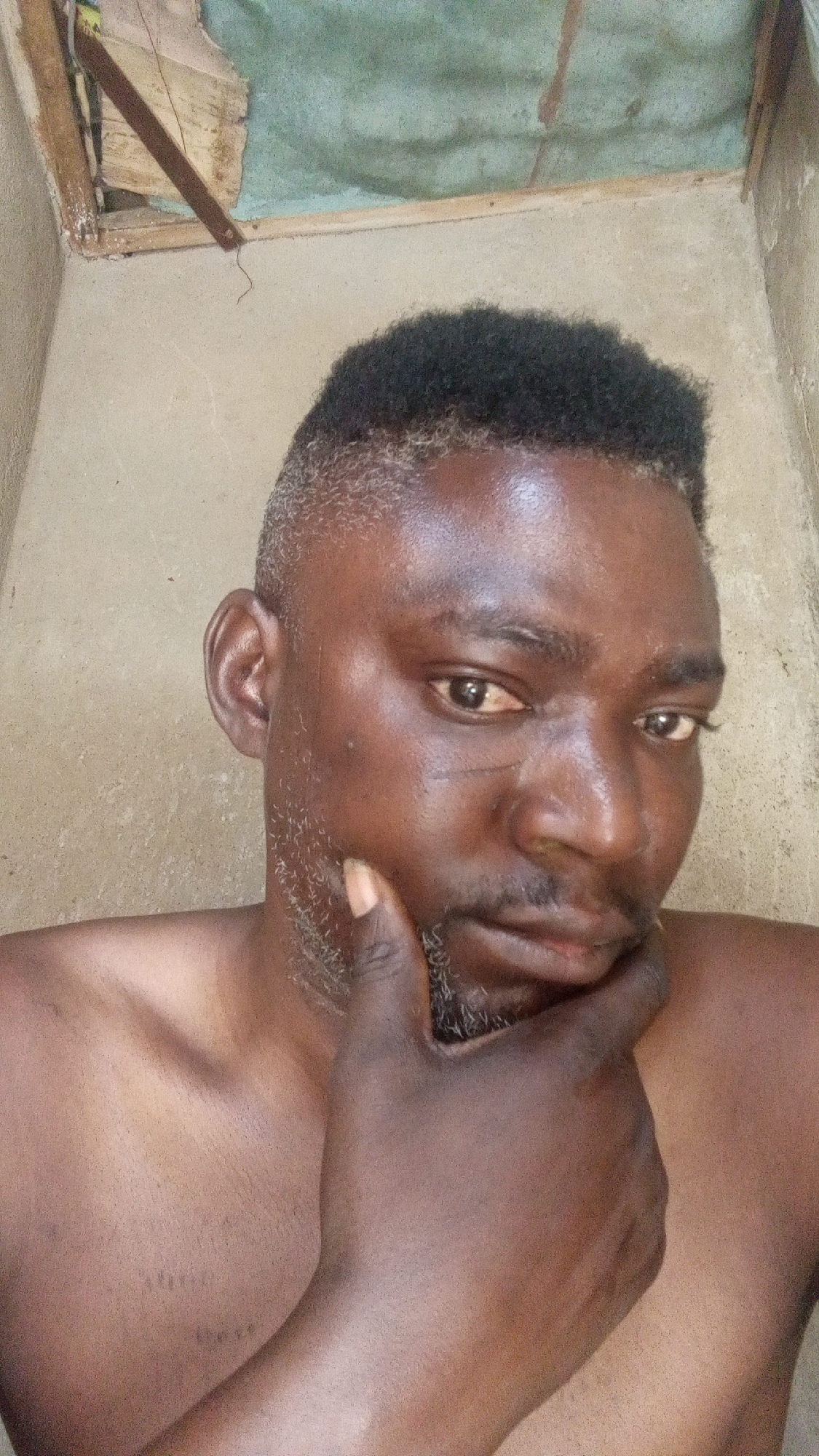 A close-up selfie shows an adult Black man from the chest up, positioned slightly to the right of the frame, looking directly at the camera. He has short, dark, textured hair with faded sides that show some graying, particularly above his left ear. His face features a short beard or stubble, also with visible gray hairs, and a scar is visible on his left cheek, running diagonally from below his eye towards his nose. He has dark eyes and a neutral to slightly contemplative expression. His right hand is raised, with his index finger and thumb touching his chin, while the rest of his fingers are curled under his jawline. He is shirtless, revealing his chest and shoulders.
The background appears to be an interior or semi-enclosed space with rough, light-colored walls, possibly concrete or unfinished plaster. Above his head, the ceiling shows signs of being unfinished or damaged, with exposed wooden supports and a section of green material, possibly mold or a painted surface. A thin, dark brown string or strap hangs down from the ceiling structure near the top left. The lighting is natural, highlighting his skin. No text is visible in the image.