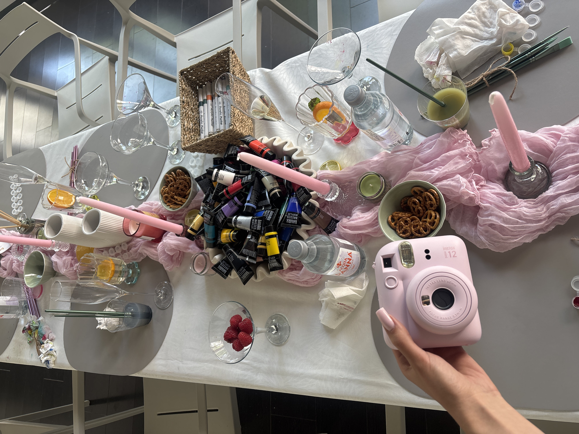 The image depicts a close-up, overhead view of a table setting, likely for an art-themed gathering or celebration. The table is covered with a white cloth, and several gray placemats are visible.
Prominently featured in the foreground is a pale pink Instax Mini 12 instant camera, held by a person's left hand with long, manicured fingernails. The camera is positioned at an angle, suggesting it's about to take a picture or has just taken one.
Scattered across the table are various art supplies, including tubes of acrylic paint in a disorganized pile, paintbrushes, and a woven basket containing several markers. A pink candle in a glass holder is also present, along with a pink decorative cloth draped artfully across the table.
Several glasses are on the table, some containing liquids like champagne and what appears to be green juice or a cocktail with mint. There are also cocktail glasses, some empty and others with remnants of red liquid. A glass bowl holds pretzels.
Other objects on the table include a bottle of Acqua Panna water, napkins, and a slice of orange in one of the glasses. A single raspberry in a martini glass adds a touch of color. The overall impression is one of a creative and social event. The scene does not provide any specific clues to its location in Sofia, Bulgaria.