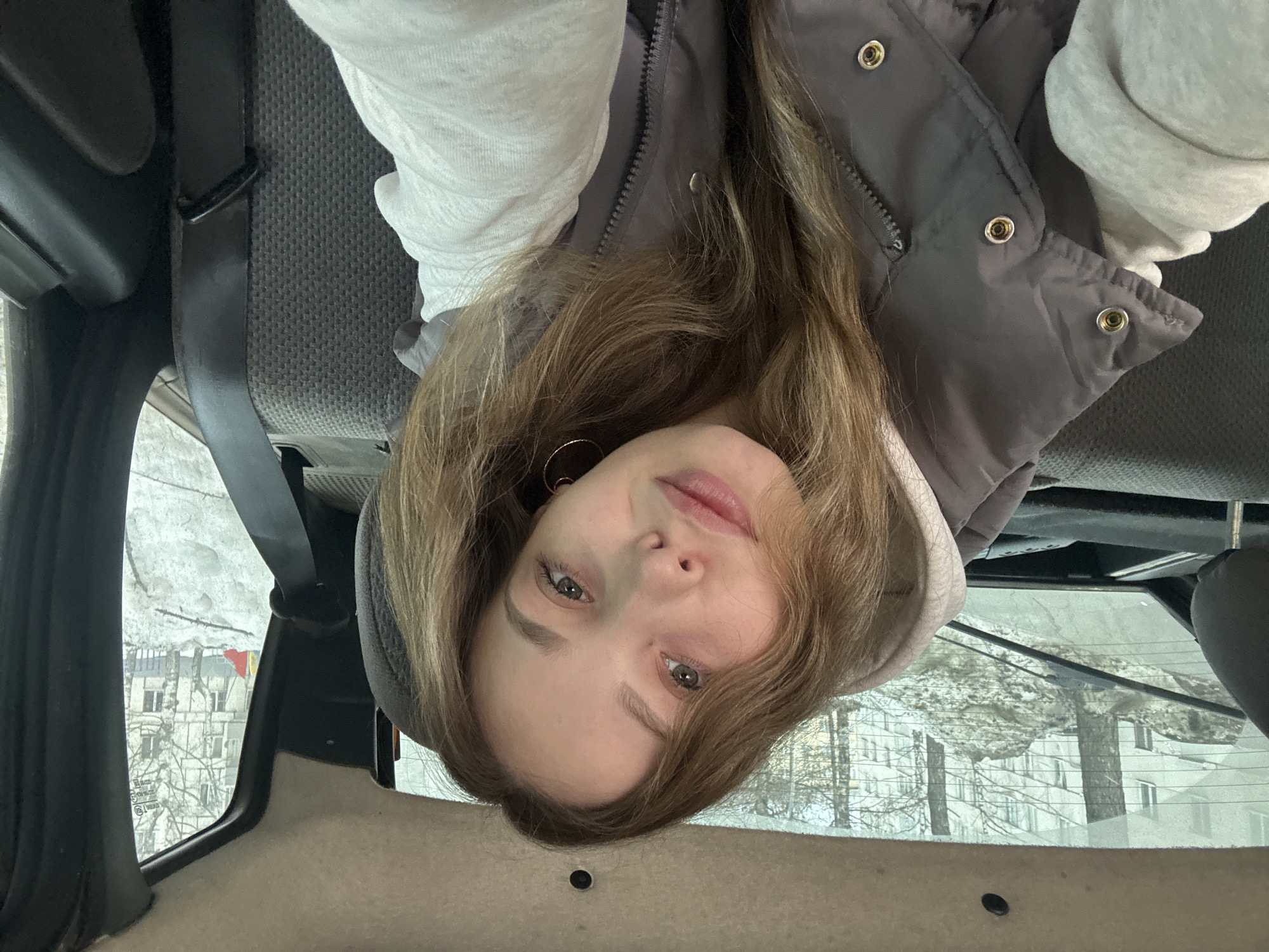 A young woman is inside a car, likely in the backseat. She is wearing a light grey hooded sweatshirt and a dark grey puffer vest. She has long, light brown hair and fair skin. The car's interior is visible, including the seat, seatbelt, and window. Outside the window, a building and trees are visible, with a hint of snow on the ground. The image is taken from an unusual angle, with the camera tilted upside down. The location is Izhevsk, Russia.
