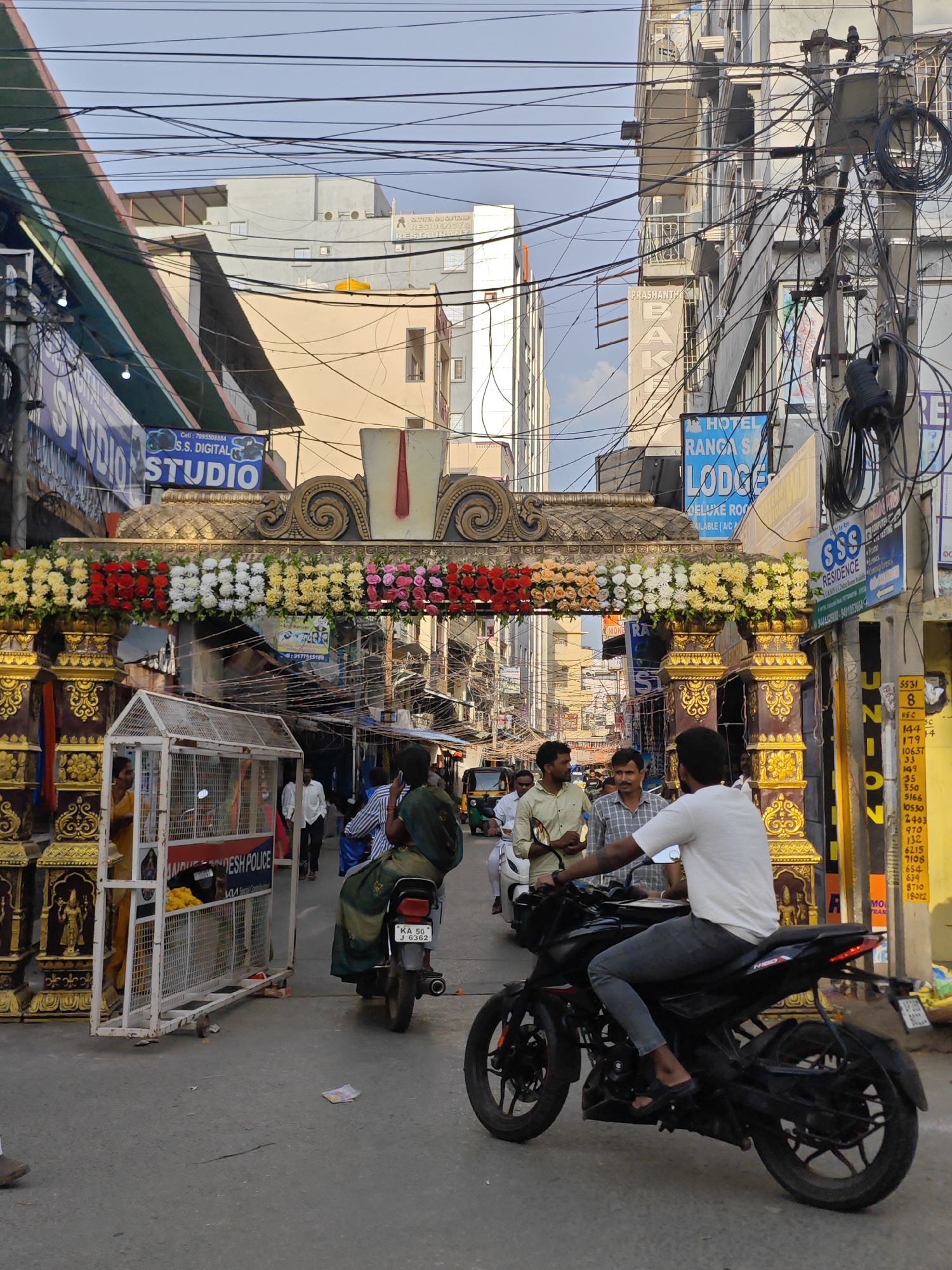 The image shows a street scene in Puttaparthi, India. In the foreground, a man on a black motorcycle is riding down the street. He is wearing a white t-shirt and grey pants. Behind him, a woman is riding on the back of a motorcycle, wearing a green saree. The motorcycle has a license plate that reads "KA 50 J 6362".
Further down the street, there are several buildings on either side, with a lot of electrical wires overhead. There are also shops and signs visible, including "HOTEL RANGA SAI LODGE" and "SSS RESIDENCE". A decorative archway adorned with flowers spans the street, with what appears to be a police barricade underneath it. A few people are walking around the area, and a three-wheeled auto-rickshaw is visible in the distance. The sky is partly cloudy, suggesting it is daytime.