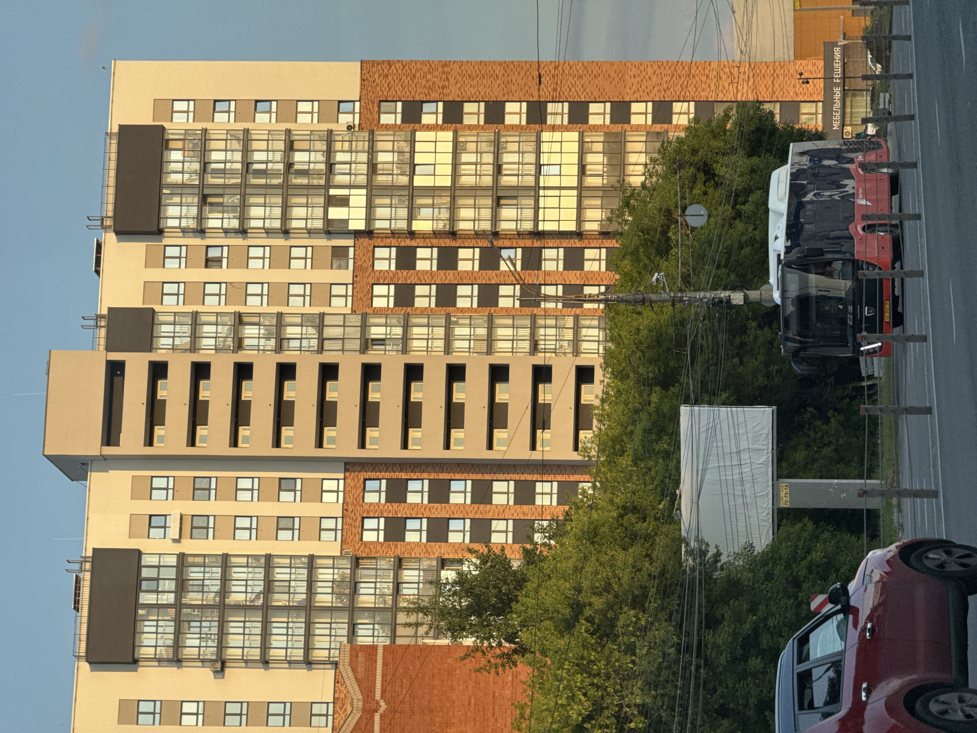 The image depicts a modern apartment building in Izhevsk, Russia, with a bus and a red car in the foreground. The building is tall, featuring a multi-colored facade with sections of light yellow, beige, and red brick. It has numerous windows and balconies, many of which are covered with reflective glass, suggesting an urban environment.
In front of the building, a bus with a red lower half and a dark upper half is stopped at a bus stop. The bus has a graphic of a face on its side. A red car is visible to the left of the bus, partially obscuring the view of the lower portion of the building. Trees line the street, partially obscuring a billboard with a white surface behind the bus. The sky is a clear, pale blue. The overall scene suggests a typical urban streetscape during daylight hours.
