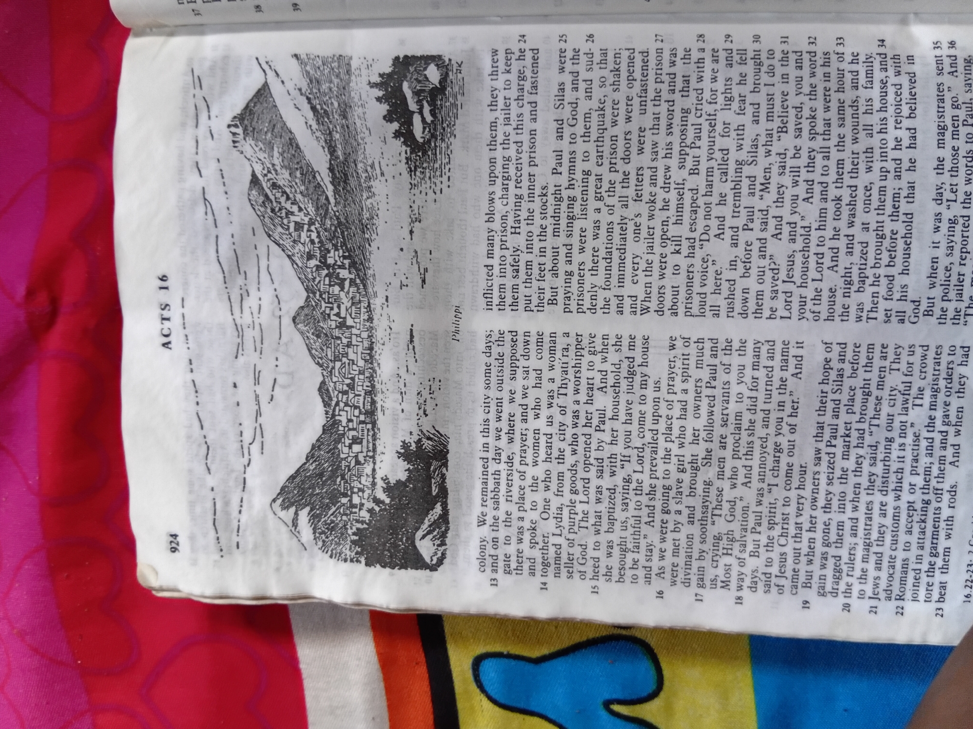 This is a photograph of an open book, appearing to be a Bible, with an illustration. The illustration depicts a cityscape nestled against a mountainous backdrop, with a prominent wall or fortification surrounding the settlement. In the foreground, a few figures are visible, suggesting a human presence within the scene.
The book's pages are white with black text. The left page has a large illustration that takes up most of the space. The right page is filled with text, organized into numbered paragraphs. Some of the text is in bold, and there are chapter and verse references. The visible text includes "ACTS 16", "Philippi", and verse numbers like "13", "14", "15", "16", "17", "18", "19", "20", "21", "22", "23", "24", "25", "26", "27", "28", "29", "30", "31", "32", "33", "34", "35", "36", "37", "38", and "39". There is also a page number "924" visible.
The page layout and the illustration suggest a historical or biblical narrative is being presented. The contrast between the detailed illustration and the dense text indicates a combination of visual and textual storytelling. The overall impression is that of an educational or devotional resource.