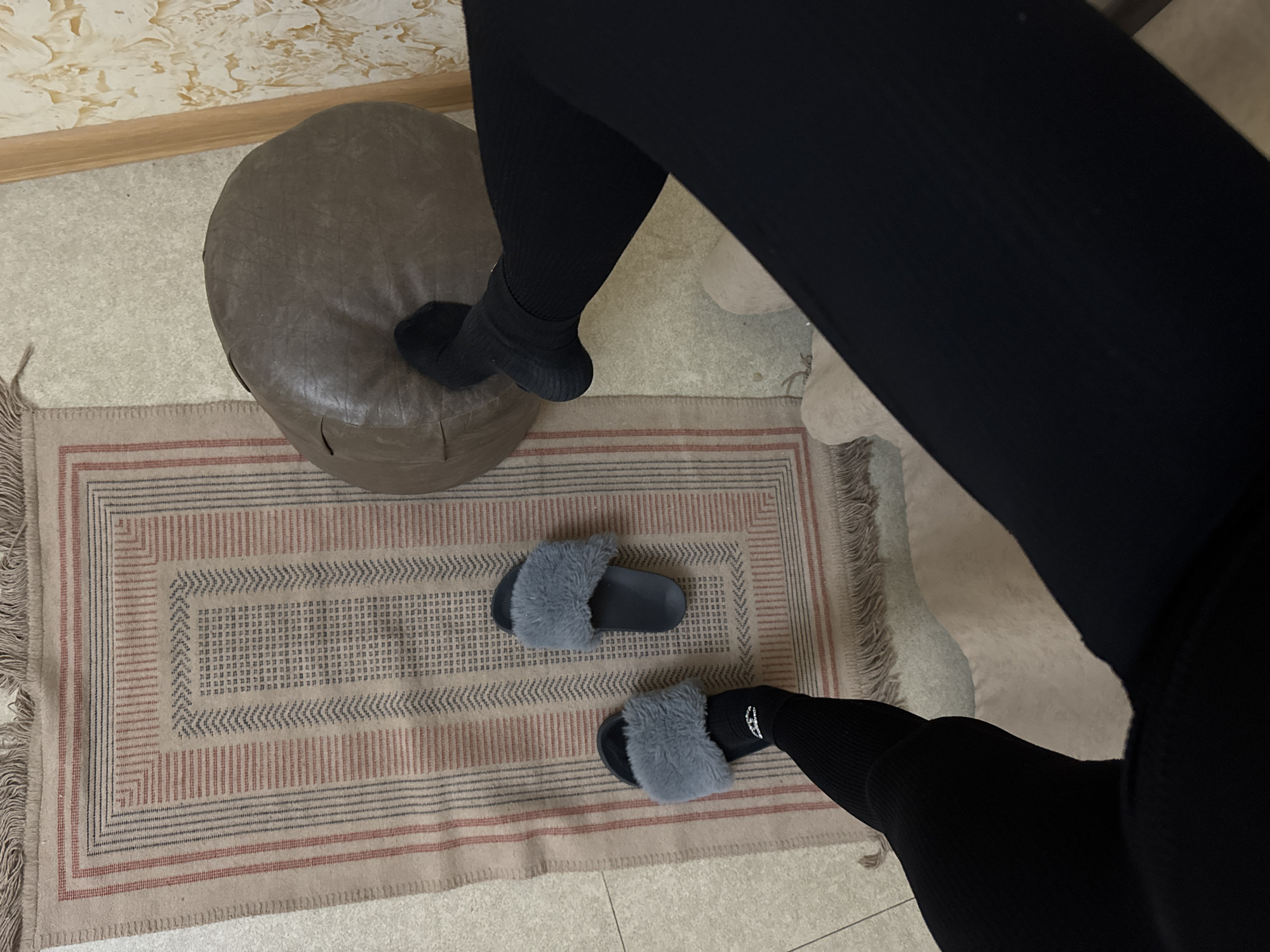 A high-angle forensic image depicts an indoor residential scene in Unknown, Unknown. The lower legs of an individual are prominent, clad in black ribbed leggings and black socks. The left foot is partially inserted into a grey, faux-fur slide sandal, while its matching sandal rests on the floor beside it. The right foot, still in a black sock, is positioned on a textured, dark brown, approximately cylindrical pouf.
A rectangular rug with a light beige base is centrally placed, displaying a geometric pattern of alternating red and dark grey thin stripes forming borders and a central field of small square motifs arranged in chevron and grid patterns. The rug features fringed ends. The floor beneath the rug appears to be light-colored tile or linoleum. To the far right, a vertical light-colored wooden trim or door frame is visible, bordering a cream-colored wall exhibiting a raised, textured, abstract relief pattern. A small segment of light beige upholstered furniture is visible in the lower right foreground.