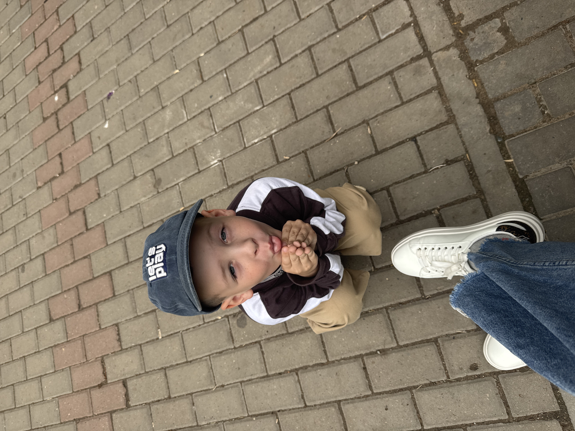 A high-angle medium shot shows a young boy wearing a dark blue baseball cap with "let's play" written in white on the front. He has his hands cupped together in front of his mouth, with his tongue slightly sticking out, and appears to be blowing a kiss. He is wearing a brown and white jacket and tan pants, and is kneeling on a gray and pink brick pathway. In the foreground, a pair of blue jeans and white sneakers are visible, suggesting the photo was taken from the perspective of the person who is photographing the boy. The environment appears to be an outdoor urban setting, possibly a park or pedestrian area.
