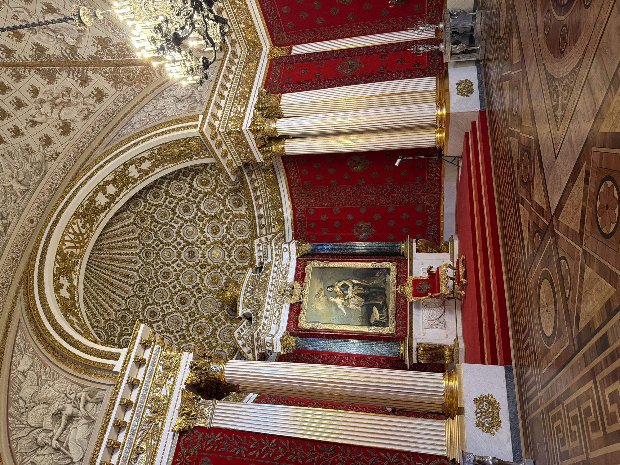 The image depicts the interior of the Field Marshals' Hall within the Winter Palace in Saint Petersburg, Russia. This opulent room is characterized by its rich red walls adorned with gold patterns and ornate gilded columns. A grand painting of two figures, possibly royalty, hangs above a red-carpeted dais. A gilded throne with a matching footstool rests on the dais. The ceiling is a masterpiece of gilded stucco work with intricate patterns and relief sculptures. A large, multi-branched chandelier hangs from the ceiling, casting a warm glow. The floor is covered with a highly detailed parquet design. In the background, there are silver decorative objects on a table and a tripod with a light fixture. The scene is illuminated by natural light and the chandelier, creating a sense of grandeur and historical significance.