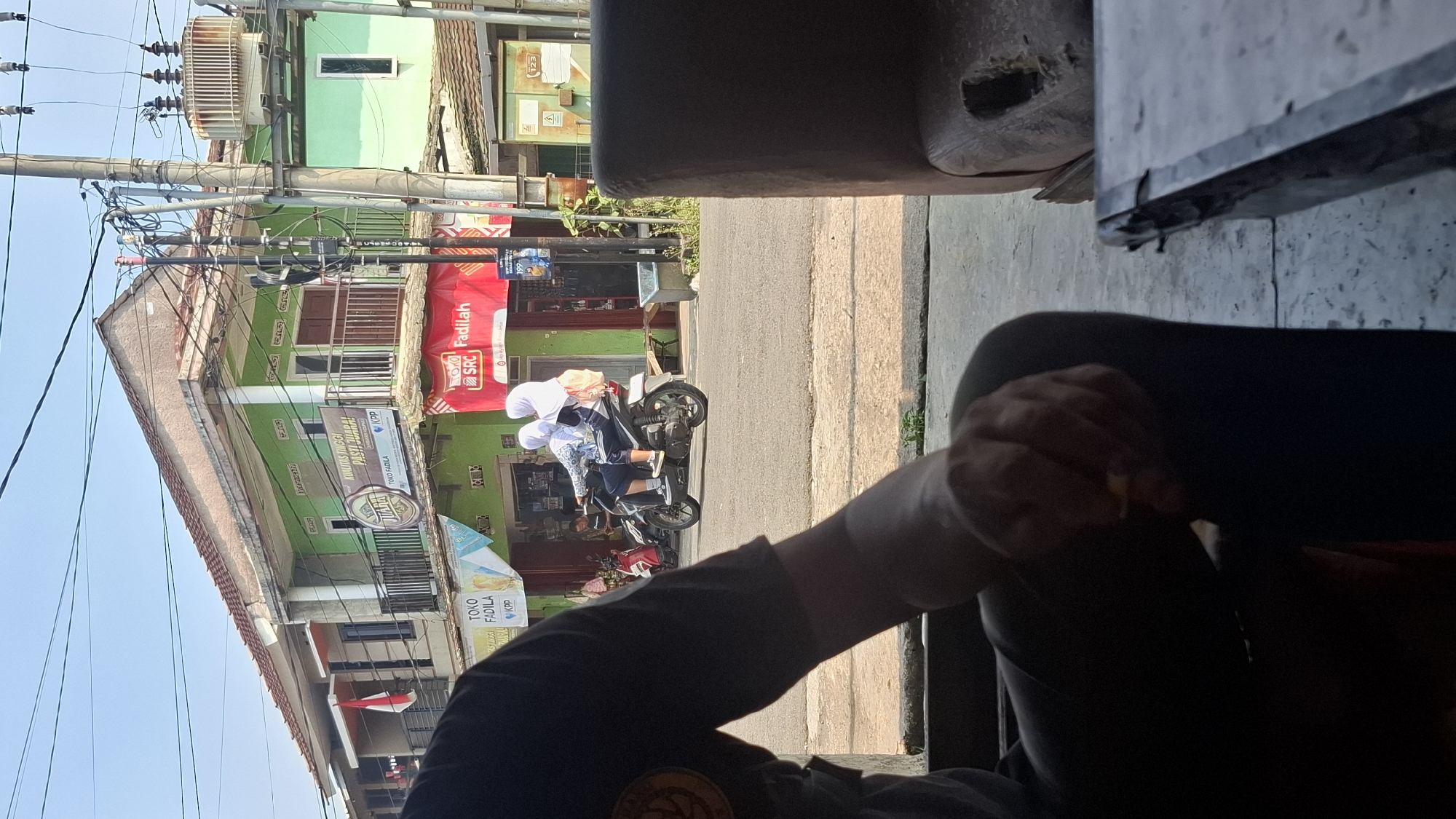 The image captures a street scene in Cilaku, Indonesia, with a focus on a building displaying various signs and an Indonesian flag. In the foreground, the dark silhouette of a person is visible, with their hand holding a cigarette. Further back, two individuals wearing hijabs ride a motorcycle on the asphalt road. The building behind them is a multi-story structure painted in green and beige, with balconies and windows. Several businesses are advertised on the building, including "TOKO FADILA" and a sign with "KUALITAS TINGGI PASTI JUARA!". Electrical poles and wires dominate the upper portion of the image, and a large electrical transformer is mounted on a pole to the right. The overall environment suggests a bustling urban or semi-urban setting with commercial activity.