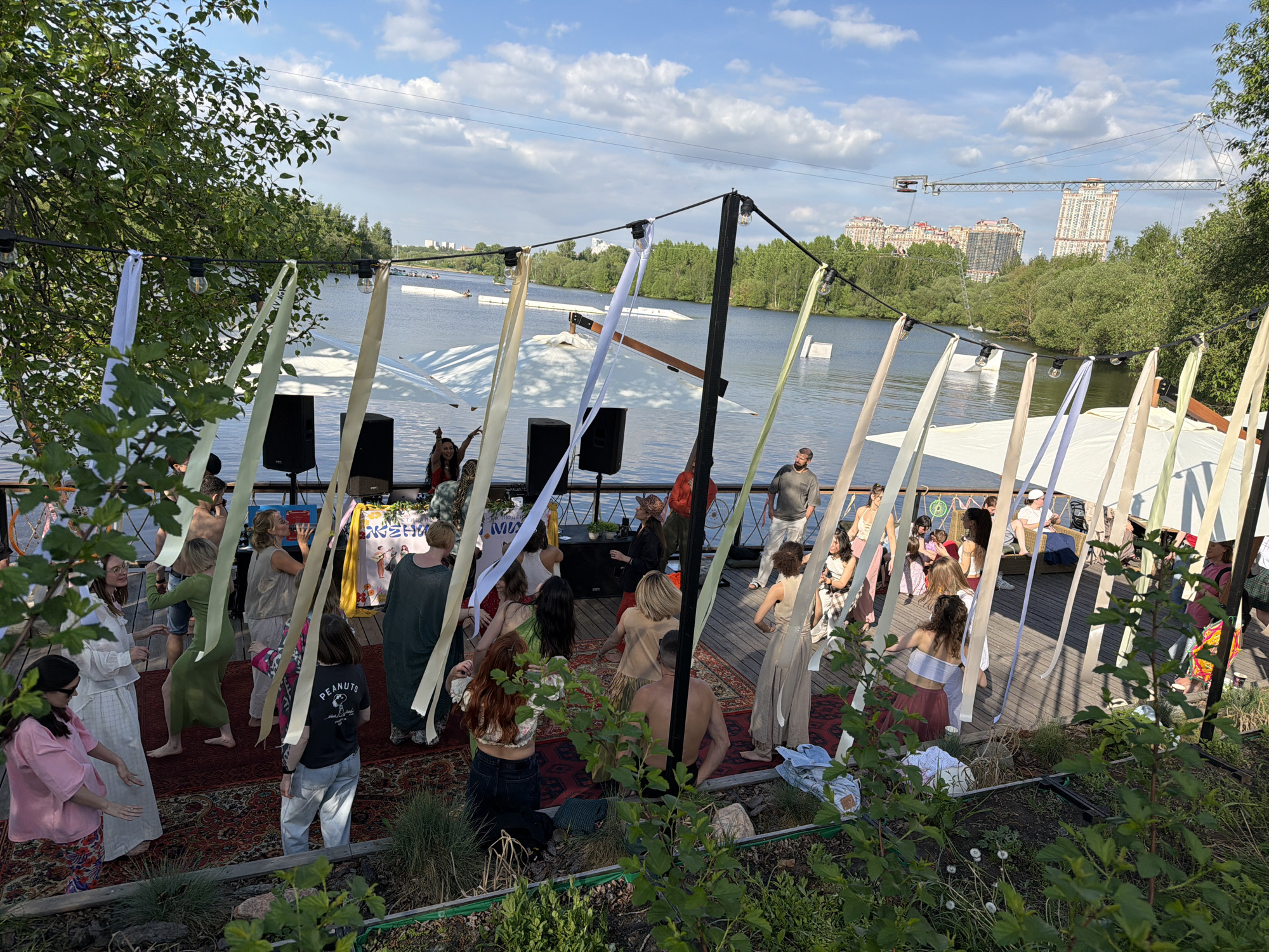 The image depicts a group of people gathered on a wooden deck by a body of water in what appears to be Khimki, Russia. The sky is partly cloudy with a mix of blue and white. The setting includes trees, a lake, and buildings in the background, some of which appear to be residential towers. A crane is also visible in the distance. A structure decorated with fabric strips is set up on the deck, under which people are congregated, some barefoot. Sound equipment is present, suggesting a possible event or gathering. A rug covers part of the deck. The deck is surrounded by greenery, including bushes. The time of day appears to be afternoon based on the lighting.