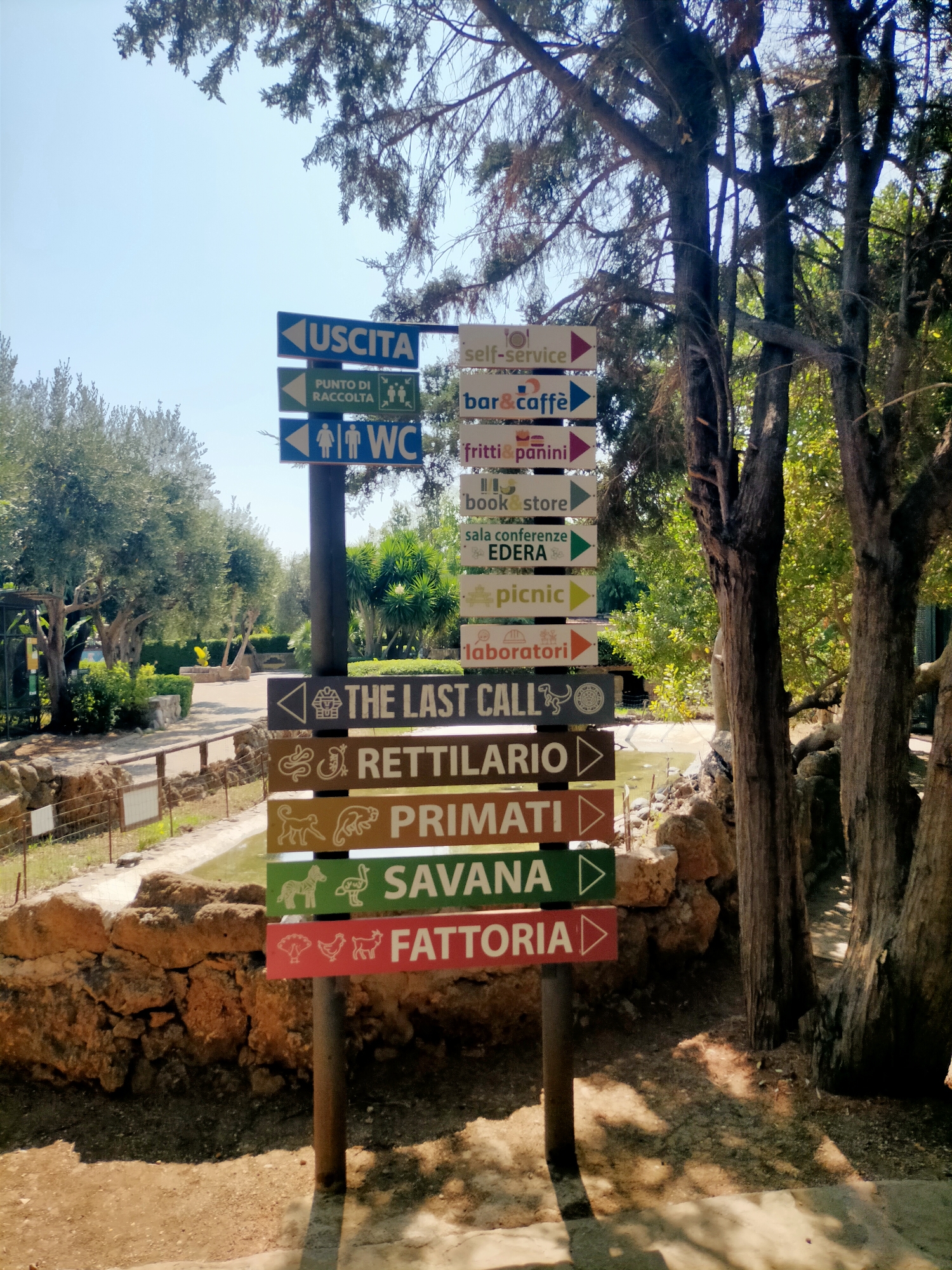 The image shows a directional signpost with multiple signs in Italian, set in an outdoor environment with trees and foliage. The signpost is made of wood and metal, with directional arrows indicating various locations. Some of the signs read "USCITA" (Exit), "PUNTO DI RACCOLTA" (Collection Point), "WC", "self-service", "bar & caffè", "fritti & panini", "book store", "sala conferenze EDERA", "picnic", "laboratori", "THE LAST CALL", "RETTILARIO" (Reptile House), "PRIMATI" (Primates), "SAVANA" (Savannah), and "FATTORIA" (Farm). There are small pictorial icons on some of the signs representing the locations. The background features trees, a path, a small body of water, and a stone wall. The sky is clear and blue. The scene appears to be in a park or zoo.