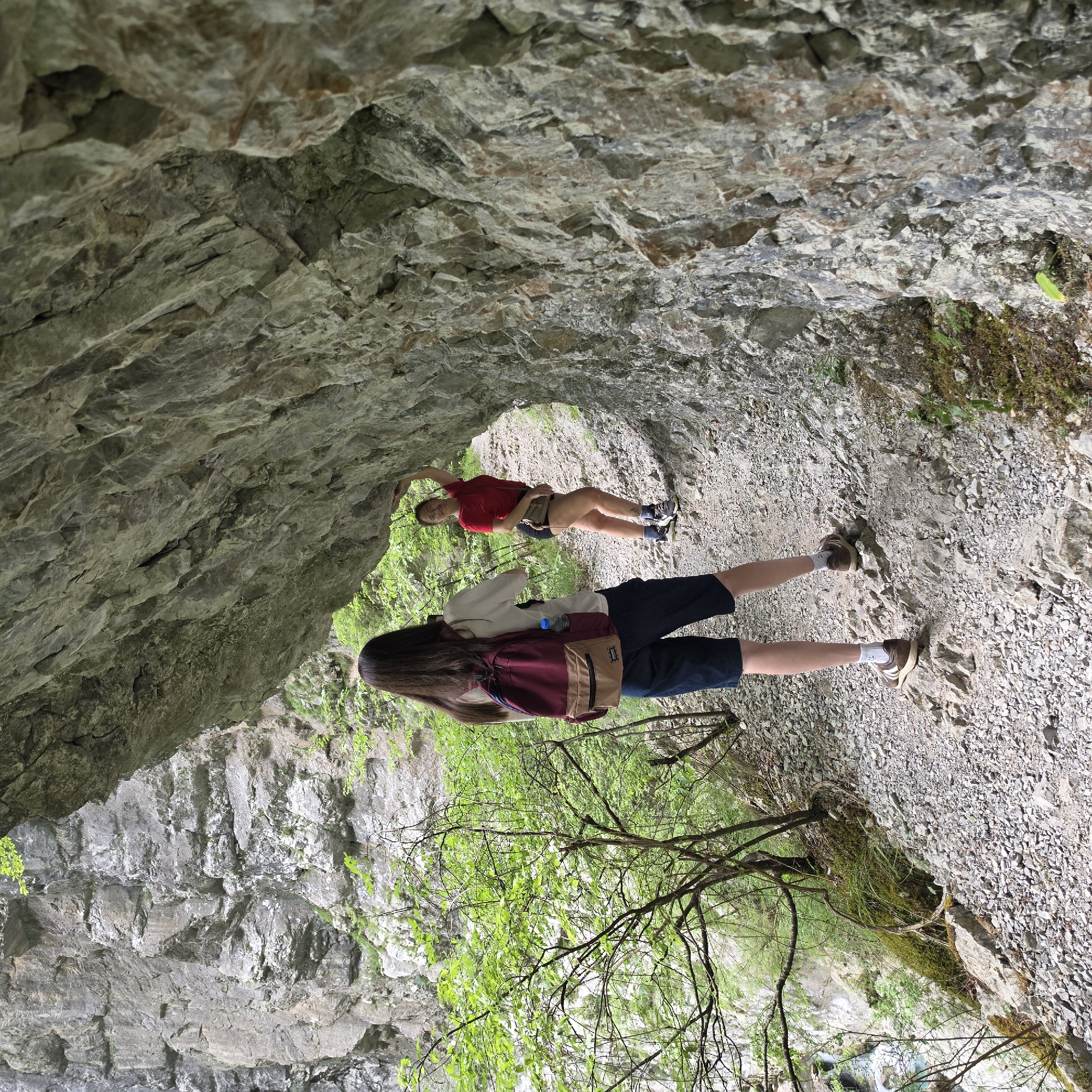Two individuals are situated within a natural rock formation in Lazba, Montenegro. One person is closer to the foreground with their back to the viewer, wearing a white shirt, dark blue shorts, and brown shoes with white socks. They are also wearing a burgundy backpack. The second person is further away, visible through an opening in the rock, wearing a red shirt and dark shorts. The environment consists of rocky terrain, vegetation, including trees and moss, and a path made of gravel and rocks. The rock formation itself is composed of layers of gray and white rock. The photograph is rotated, giving a unique perspective of the scene.