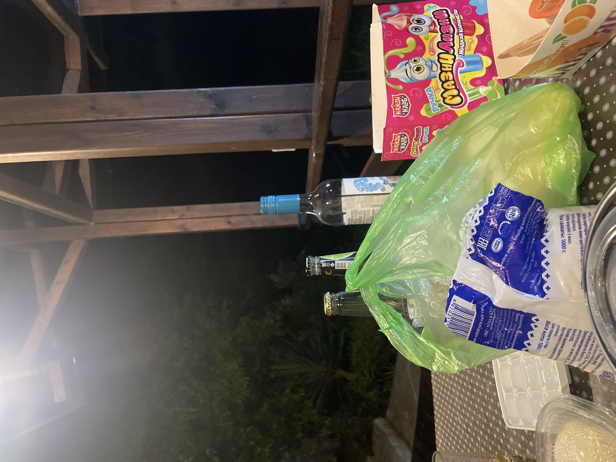 In a dimly lit outdoor setting, likely a porch or covered patio, several items are arranged on a patterned table. A bright green plastic bag dominates the foreground, obscuring much of its contents. To the left, two glass bottles, one with a gold cap and the other partially visible with a silver cap and label, are positioned near the bag. A third, taller glass bottle with a blue cap and a white label is situated to the right of the green bag. In the background, a dark, possibly wooden structure with vertical beams and horizontal planks suggests an overhead covering. Behind the table, dense foliage is visible, hinting at a natural environment. A pink cardboard box with colorful cartoon characters and text in an unfamiliar script is placed on the table, to the right of the green bag. A white plastic ice cube tray and a clear plastic container with a light-colored substance are also visible on the table. The overall scene suggests a casual gathering or aftermath of one, with remnants of beverages and packaging.